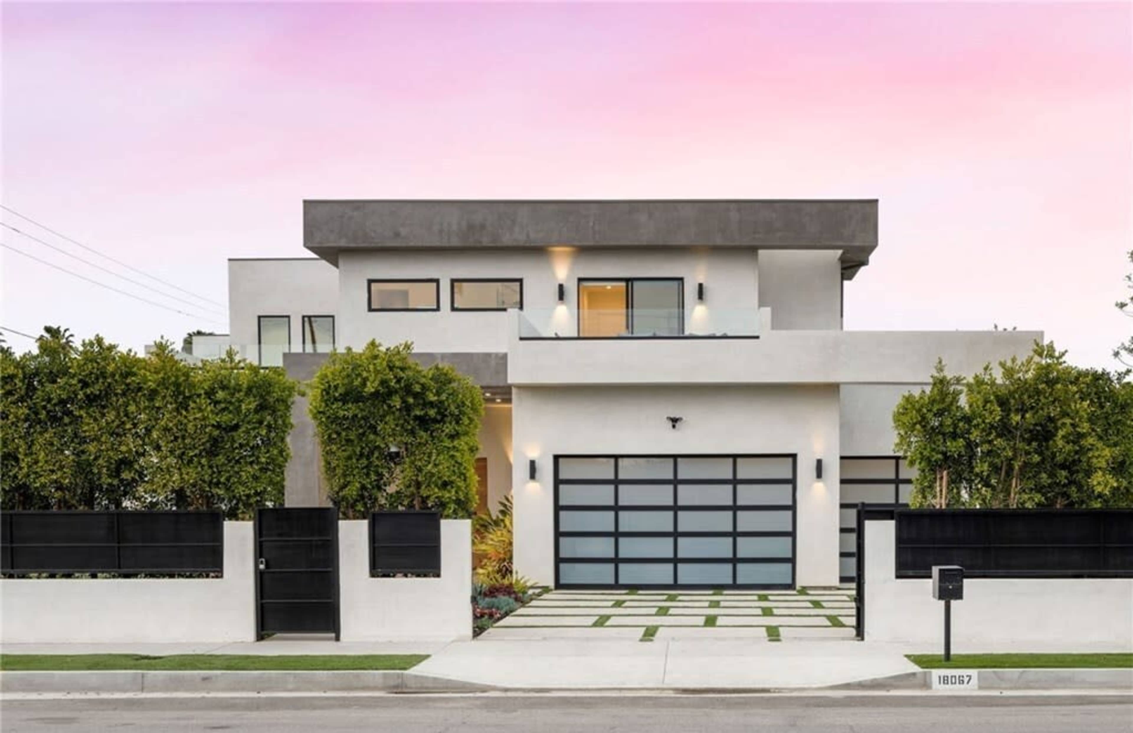 A modern two-story house with a flat roof and a large garage door is surrounded by a manicured landscape and a tall hedge.