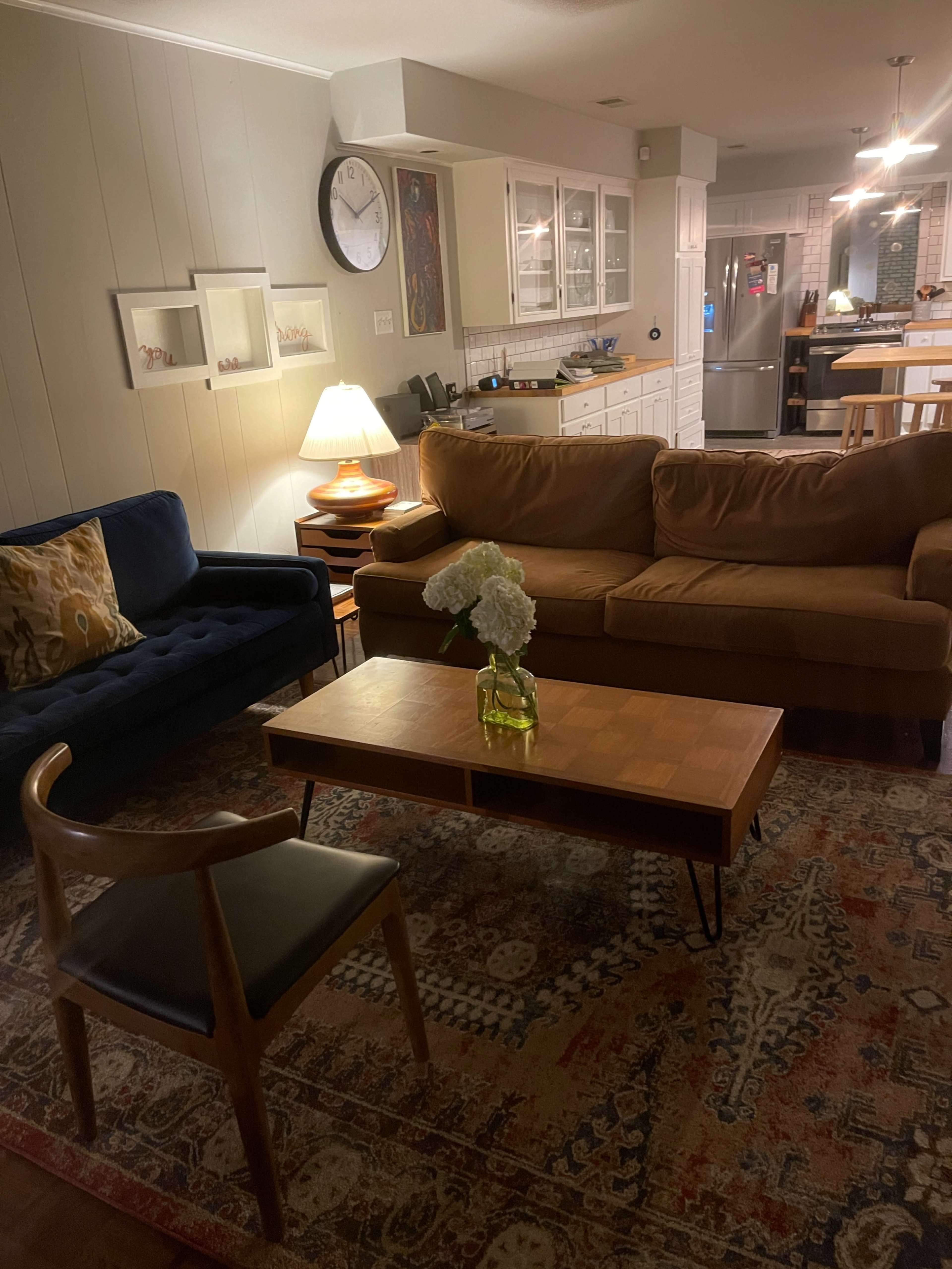 A living room features a brown sofa, a blue chair, a wooden coffee table with a vase of flowers, and a rustic area rug, with kitchen space visible in the background.