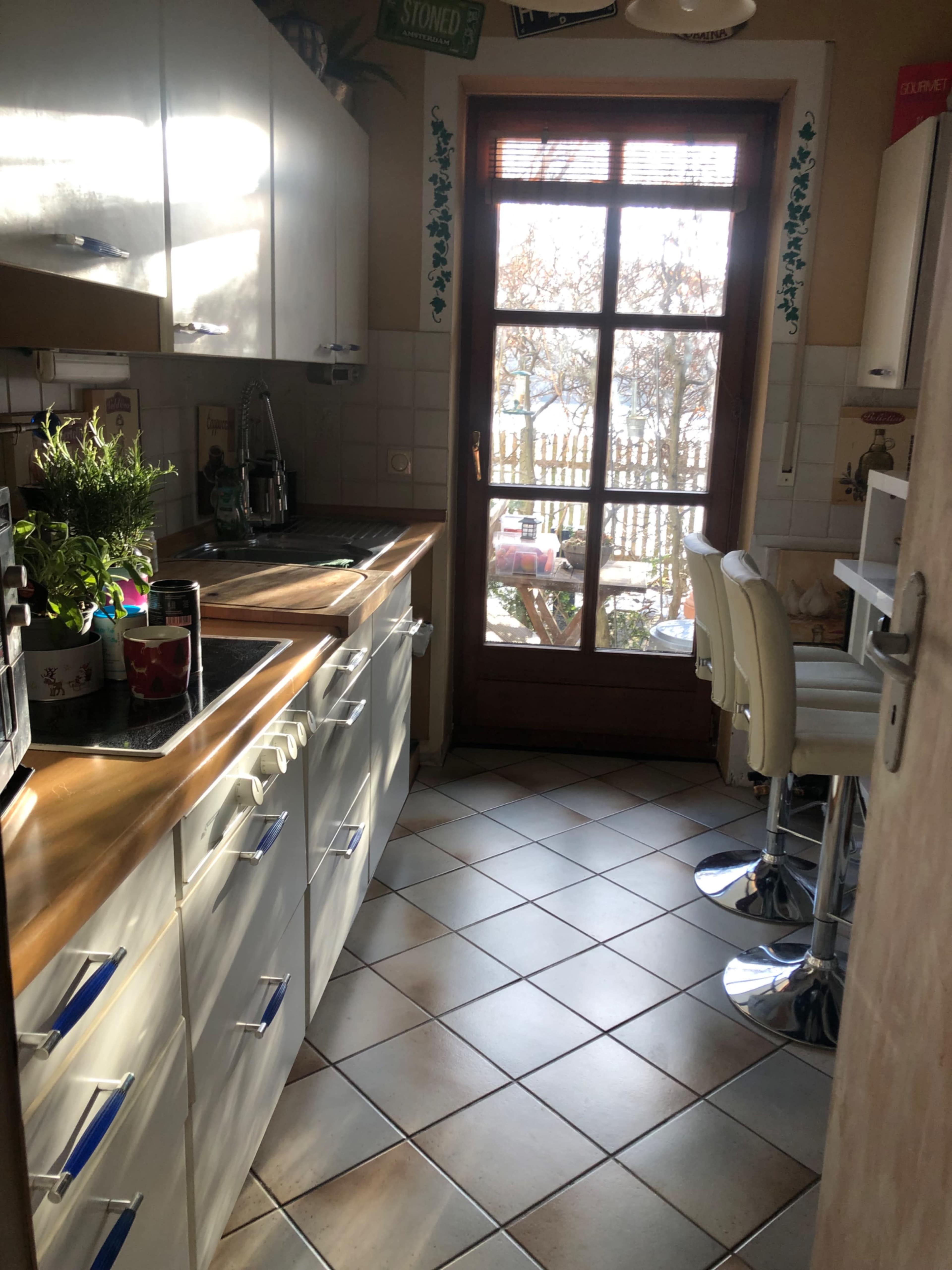 The image shows a small kitchen with white cabinets, a wooden countertop, and a door leading to an outdoor area.