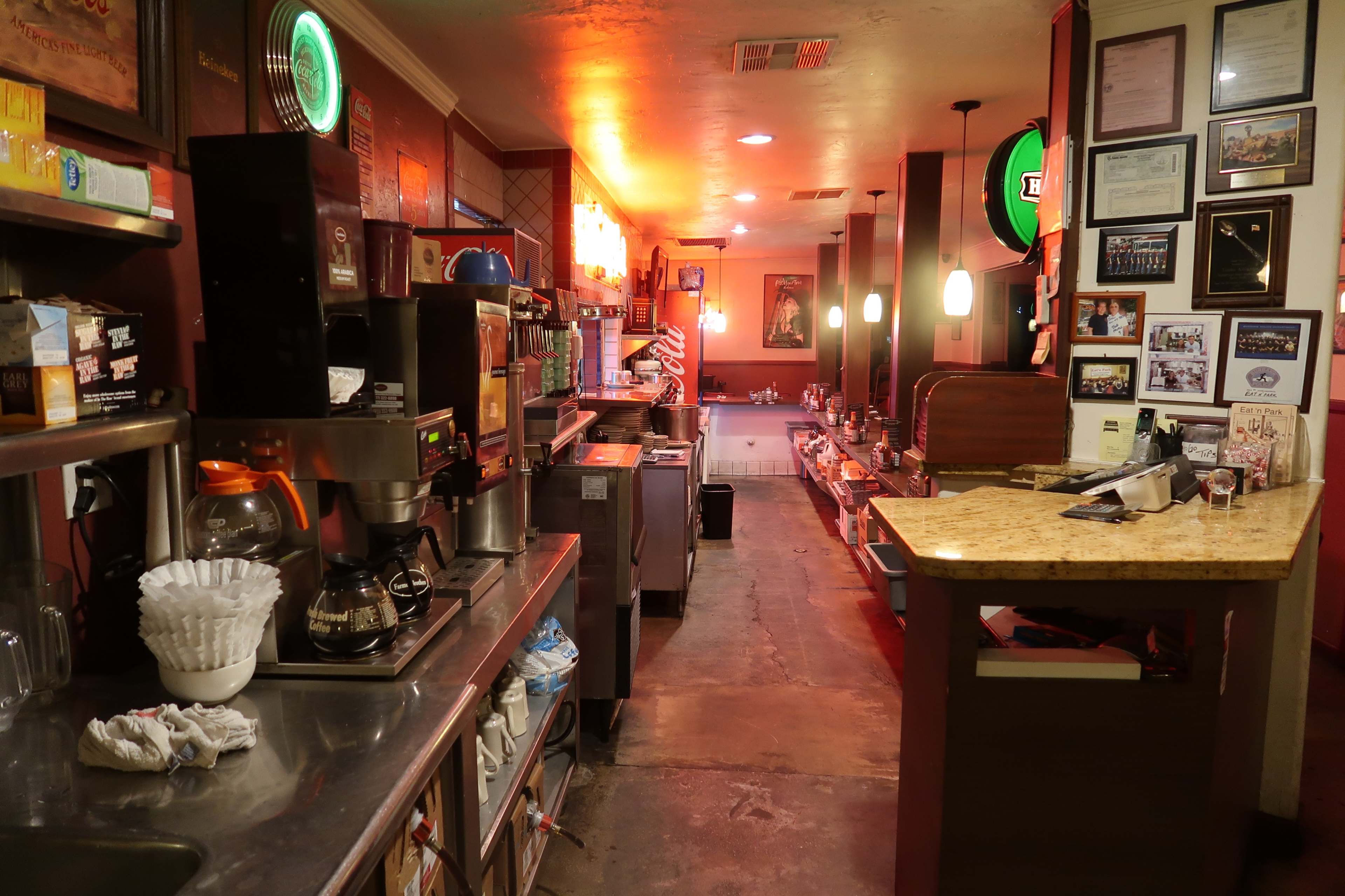 A cafe interior showcases a long counter with a coffee machine, shelves stocked with supplies, and wall decorations including framed certificates and artwork.