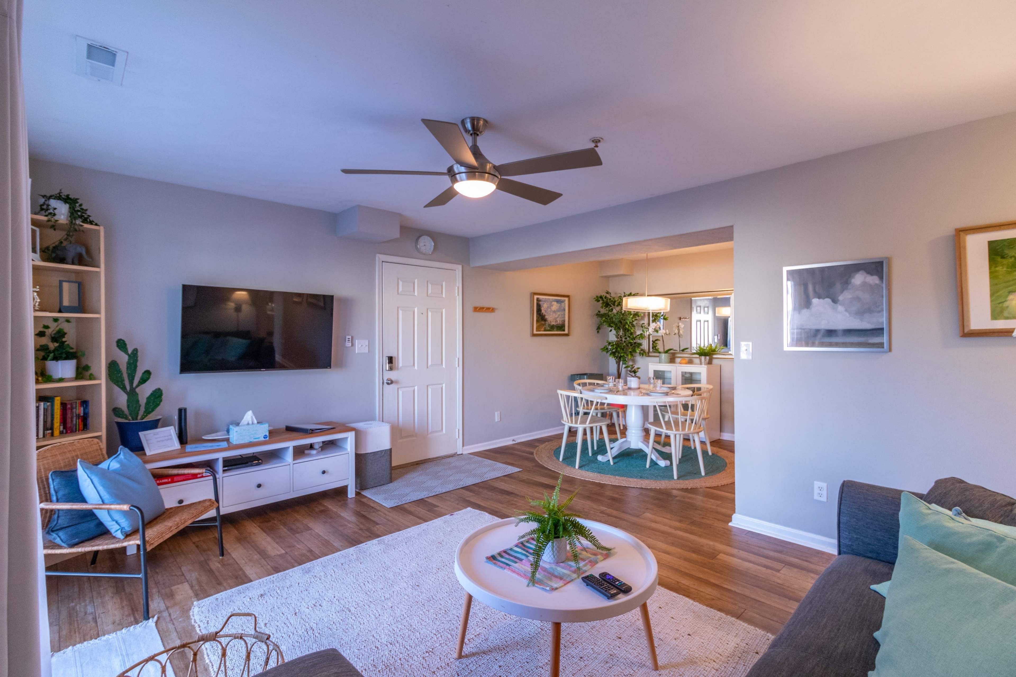A modern living room and dining area with a neutral color palette, featuring a television, a sofa, and a circular dining table surrounded by chairs.