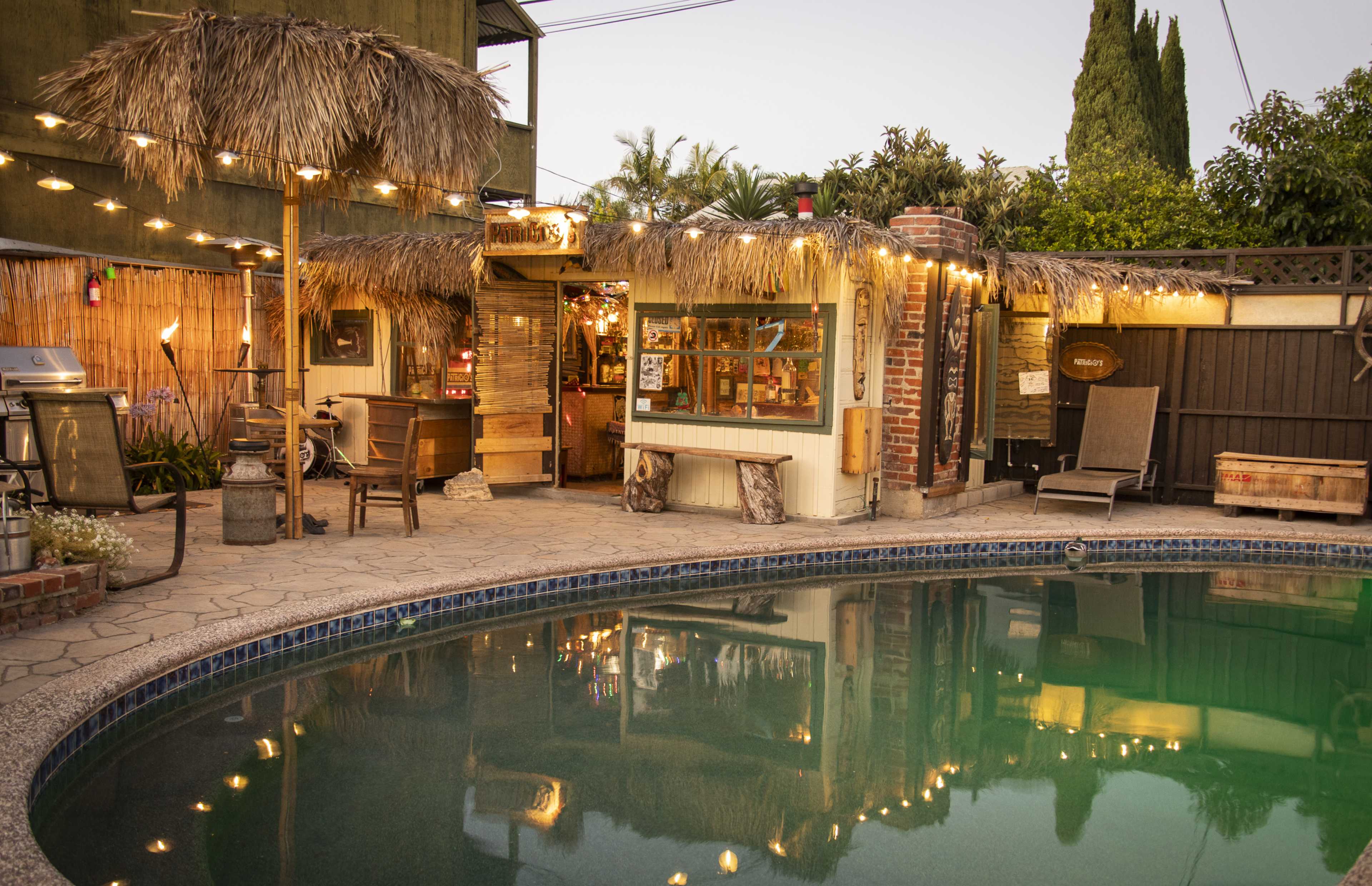 The scene features a backyard with a pool surrounded by a thatched-roof patio and string lights, creating a tropical retreat atmosphere.