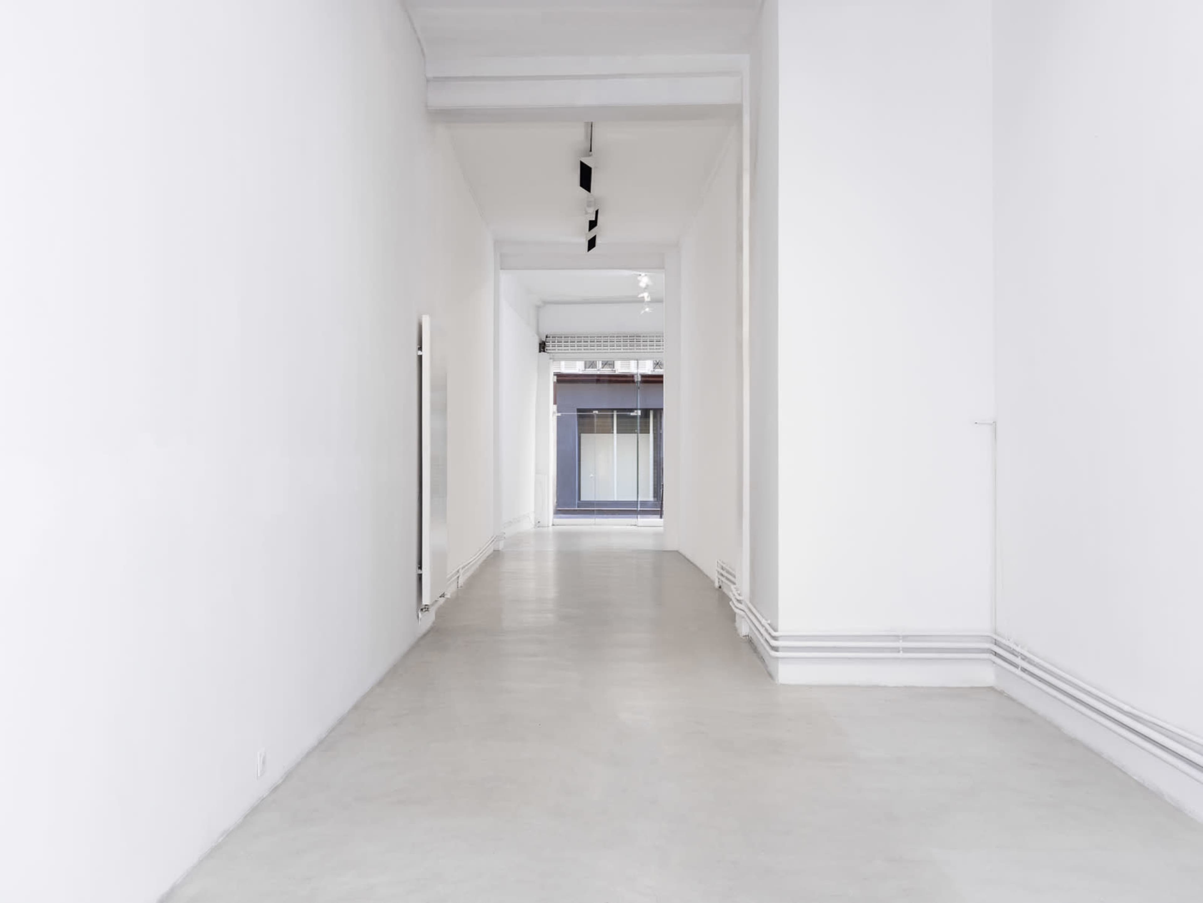 A long, empty corridor with white walls and a concrete floor leads to a doorway at the far end.