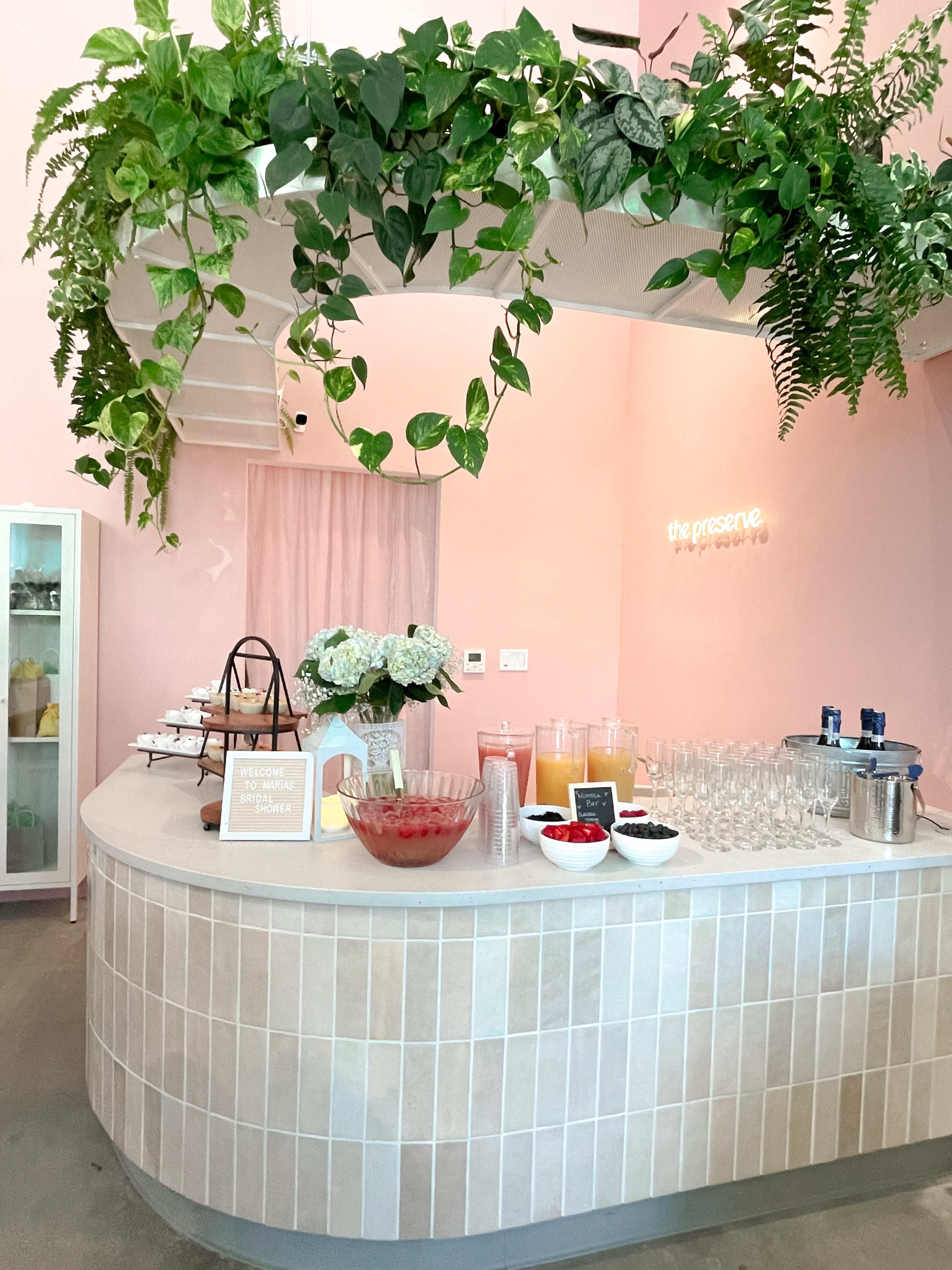 A light pink café interior featuring a curved countertop adorned with drinks and fruit, surrounded by greenery.