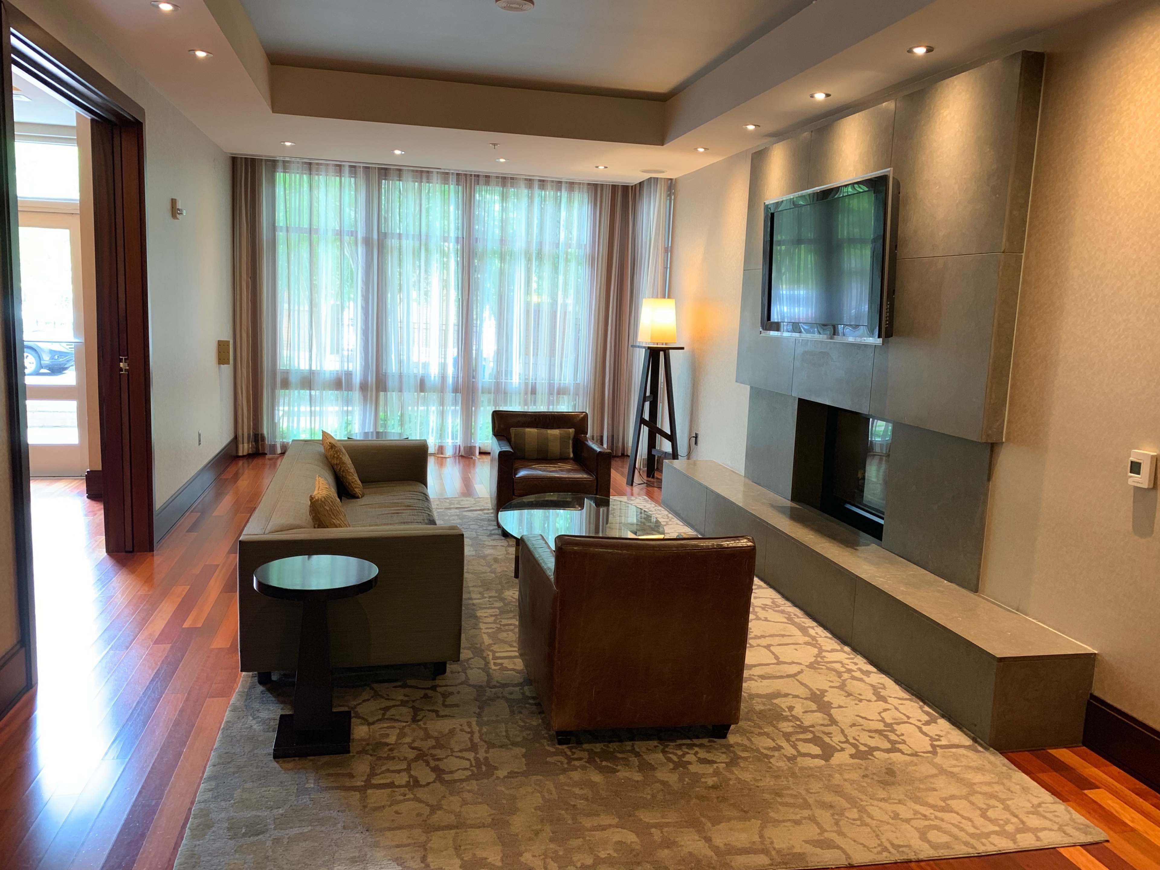 A modern living room with a flat-screen TV mounted above a stone fireplace, featuring two armchairs, a sofa, and large windows dressed in sheer curtains.