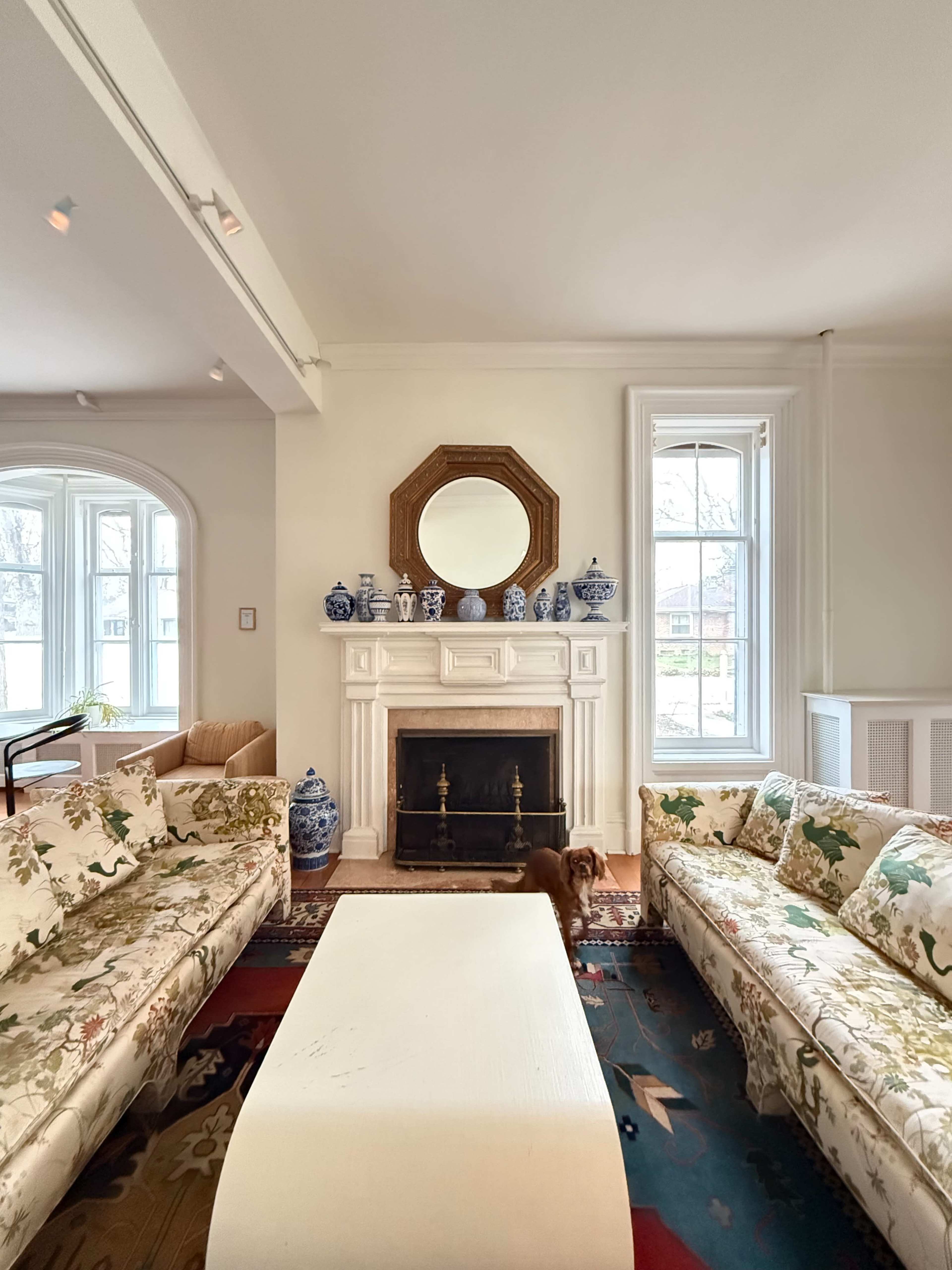The image shows a well-decorated living room featuring floral-patterned sofas, a white coffee table, a fireplace with a mirror above it, and blue-and-white ceramic decor.