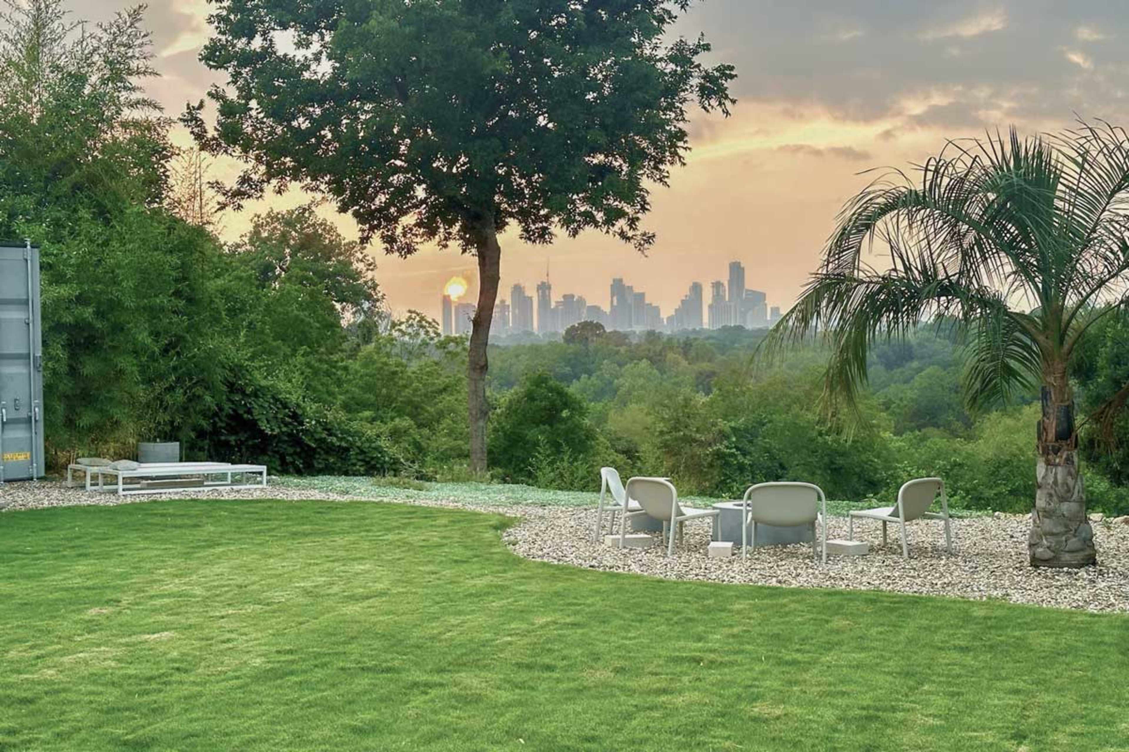 A green lawn with modern seating overlooks a city skyline at sunset, framed by trees and palm plants.