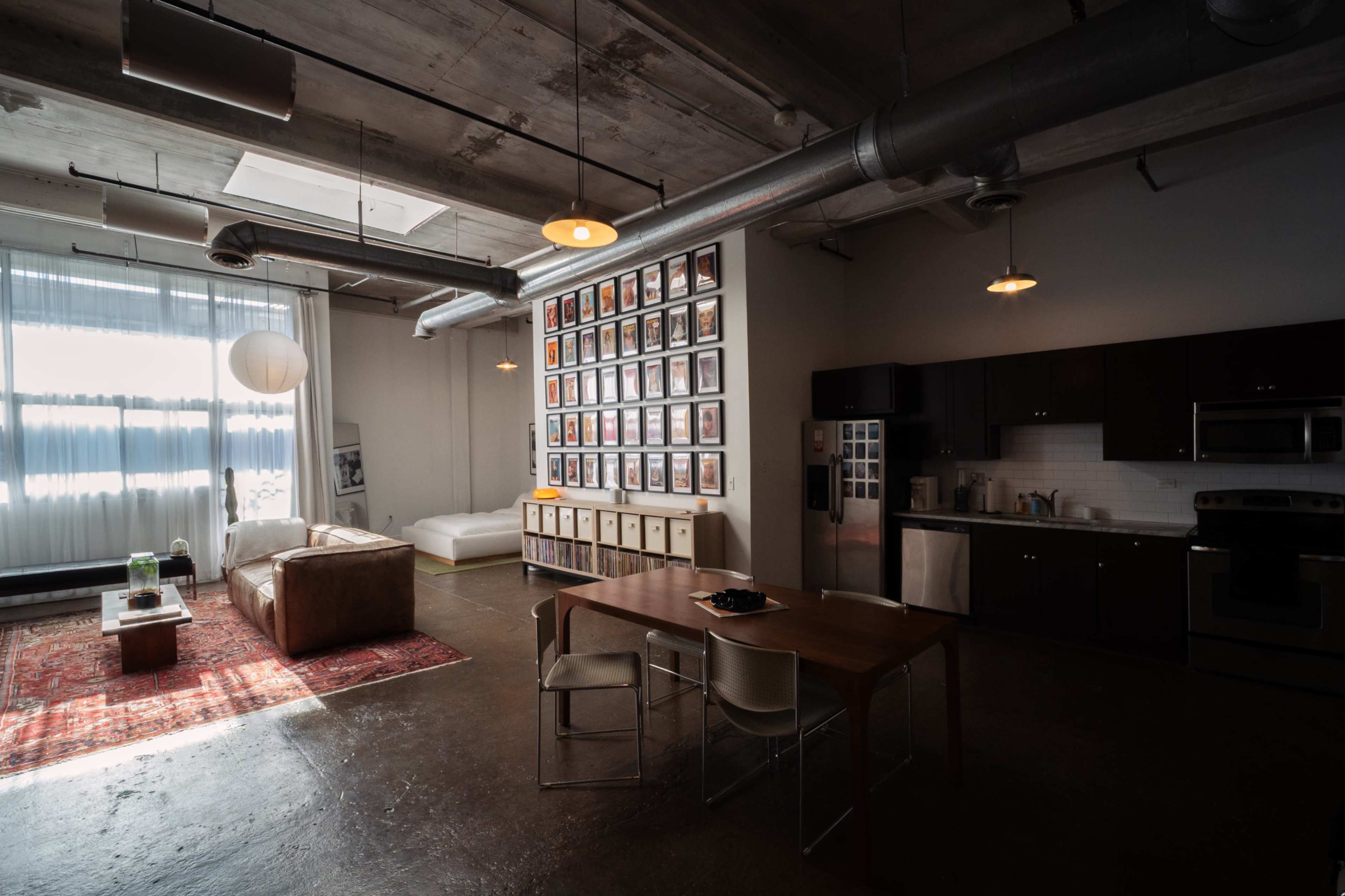 The image shows a modern, industrial-style apartment featuring a living area with a sofa and a dining table, a kitchen area, and a wall adorned with numerous framed pictures.