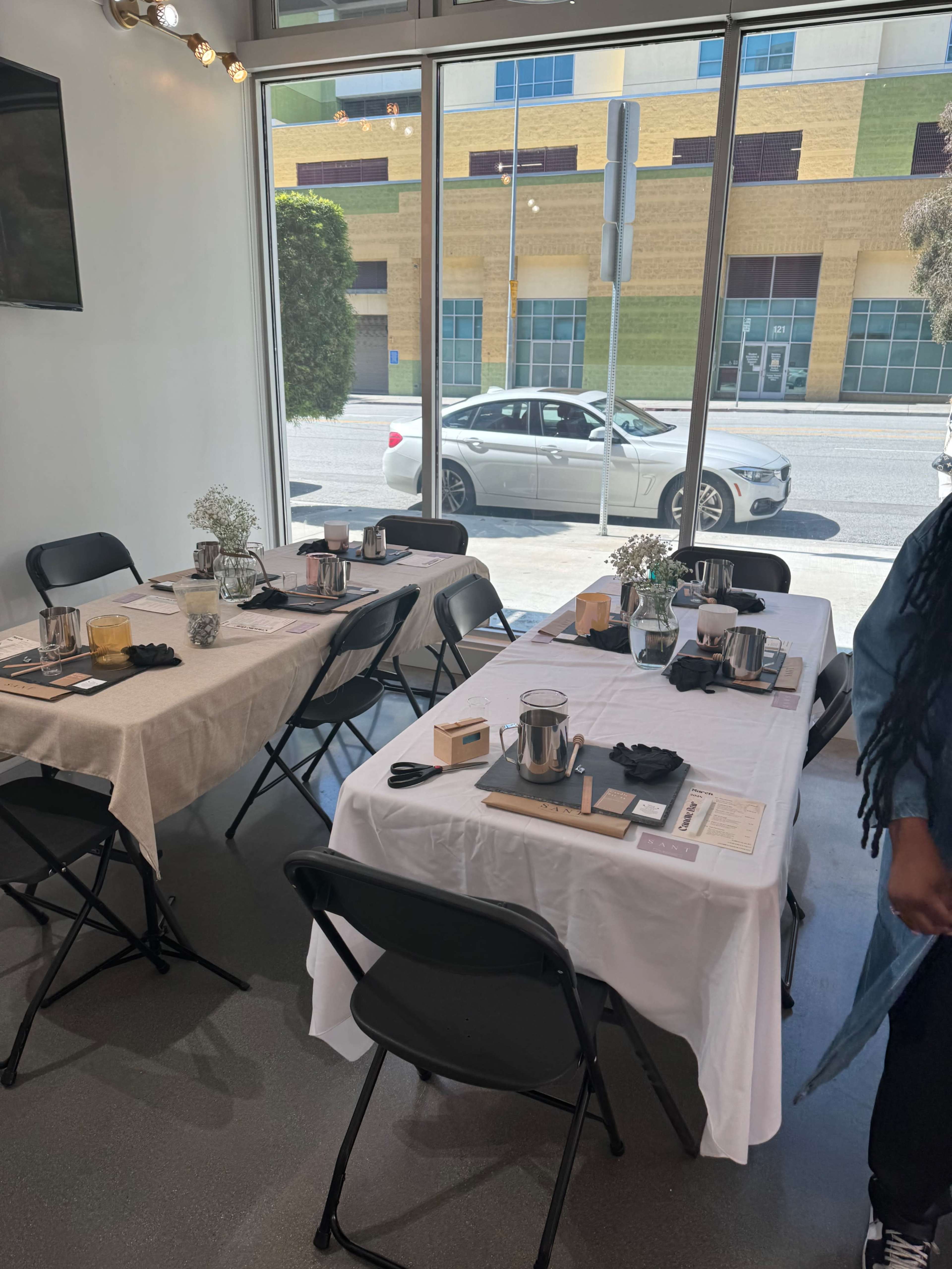 A brightly lit dining area with two tables set for a meal, featuring white tablecloths, utensils, and drinks, overlooking a street with cars outside.