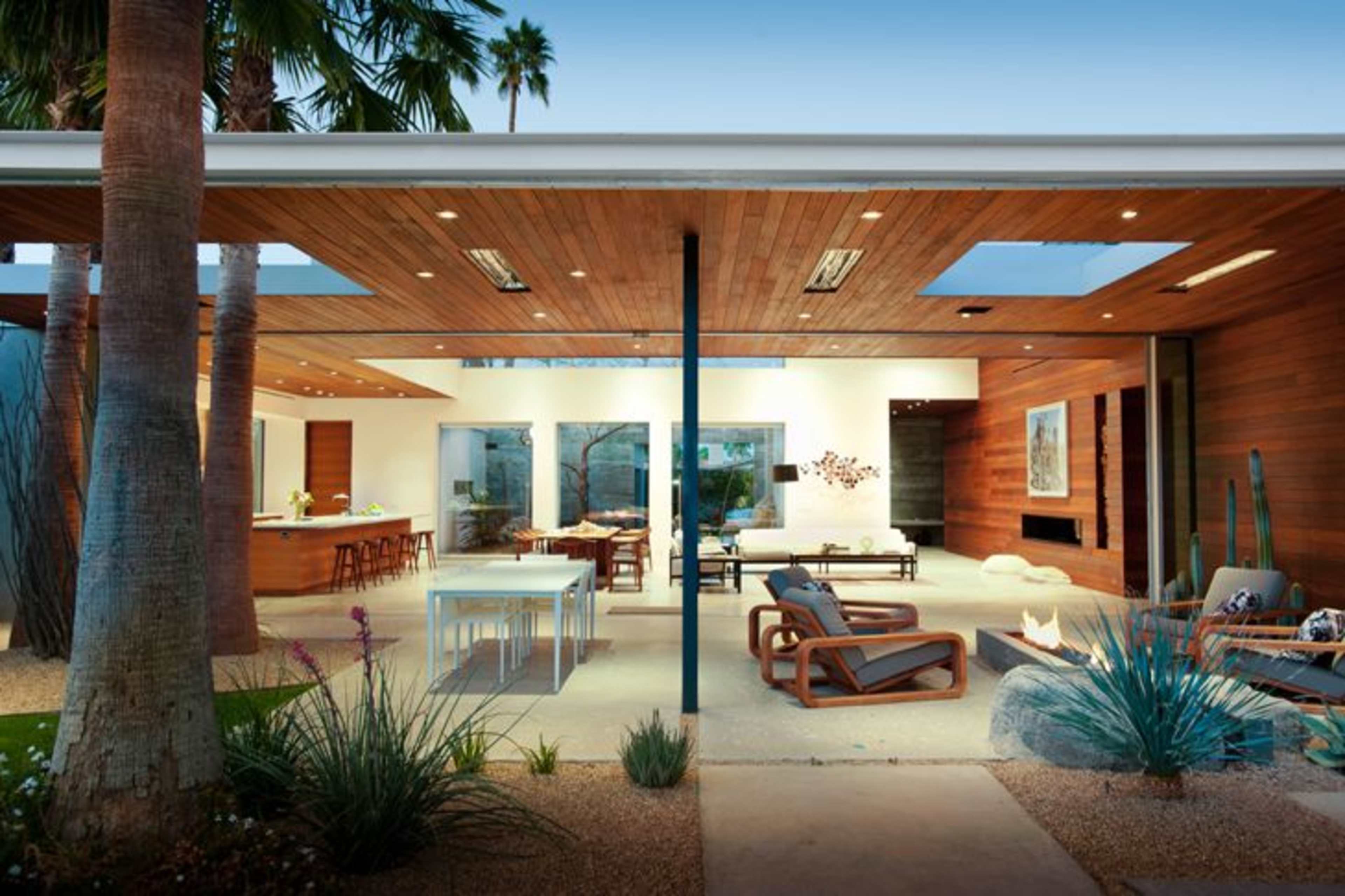 The image shows a modern outdoor living space featuring wooden accents, minimalist furniture, and lush landscaping under a clear sky.