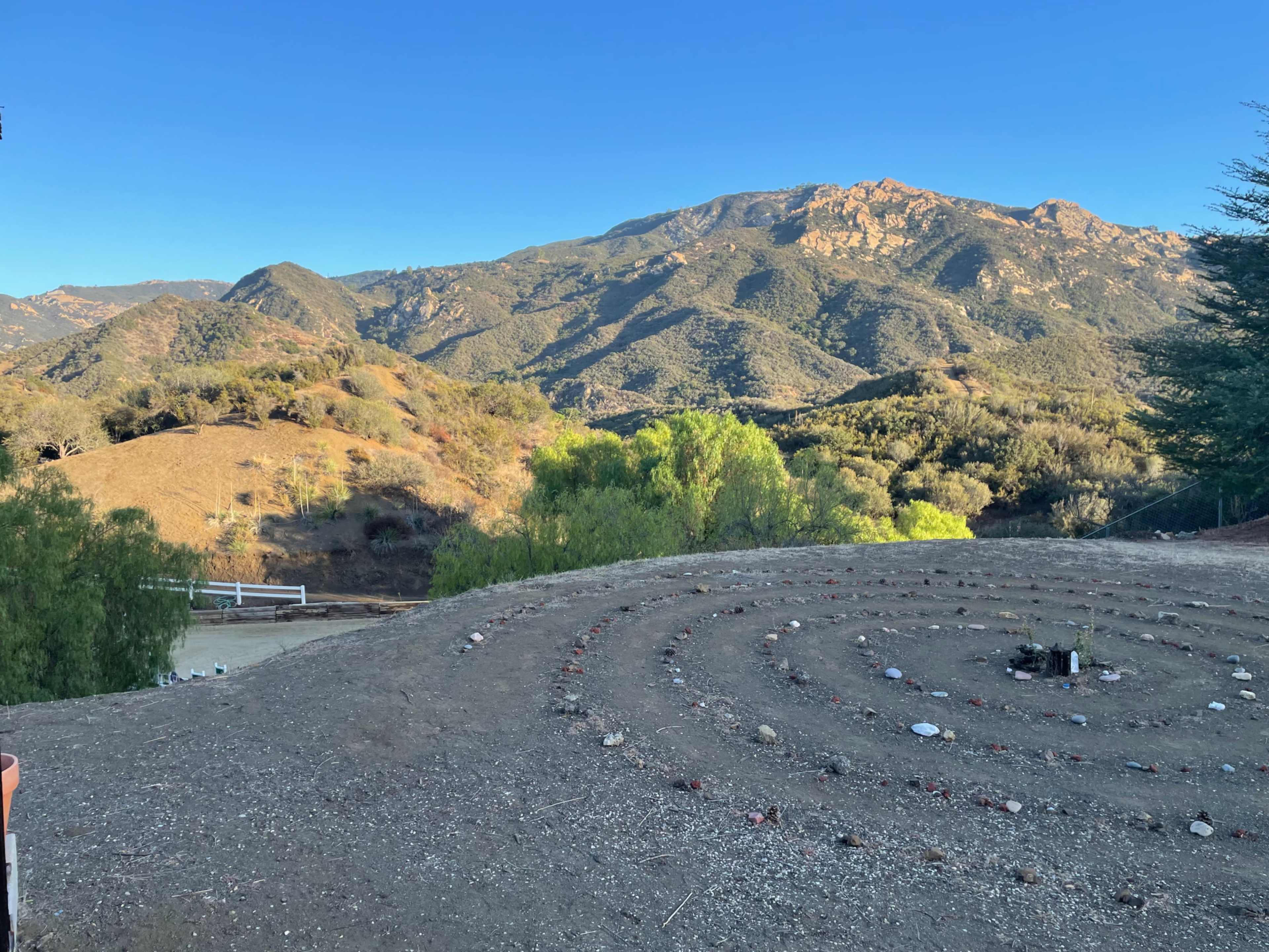 The image shows a spiral stone path in a dry landscape with rolling hills and a mountain in the background under a clear blue sky.