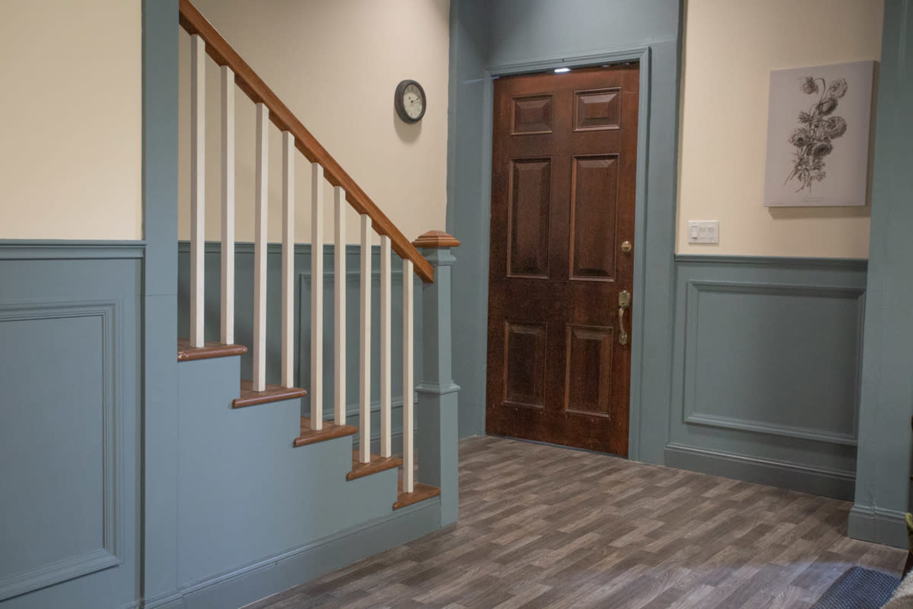 A hallway with a staircase on the left, a wooden front door on the right, and a clock mounted on the wall.