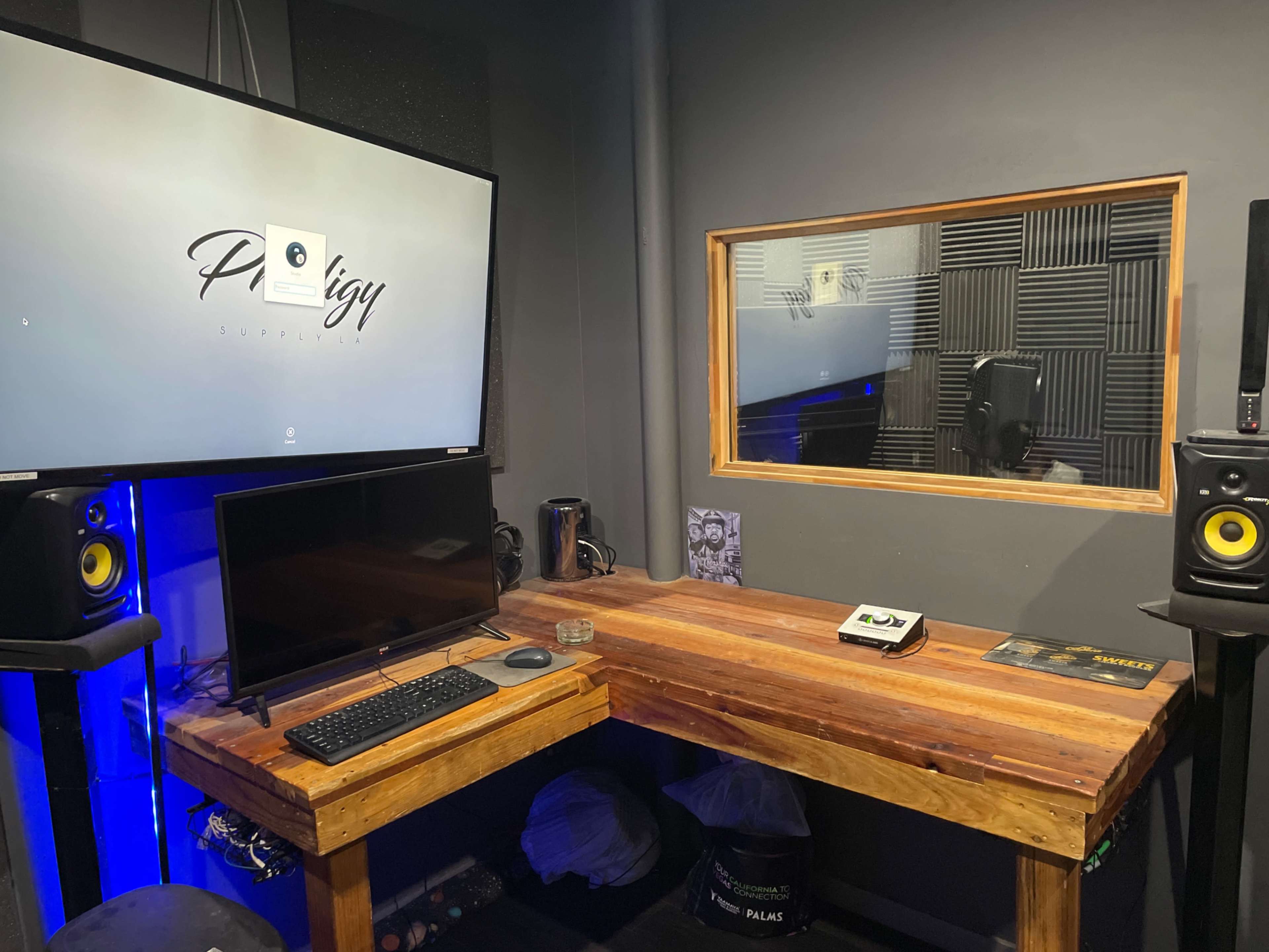 The image shows a small recording studio featuring a wooden desk with a computer, studio monitors on either side, and a large screen displaying a logo on the wall.