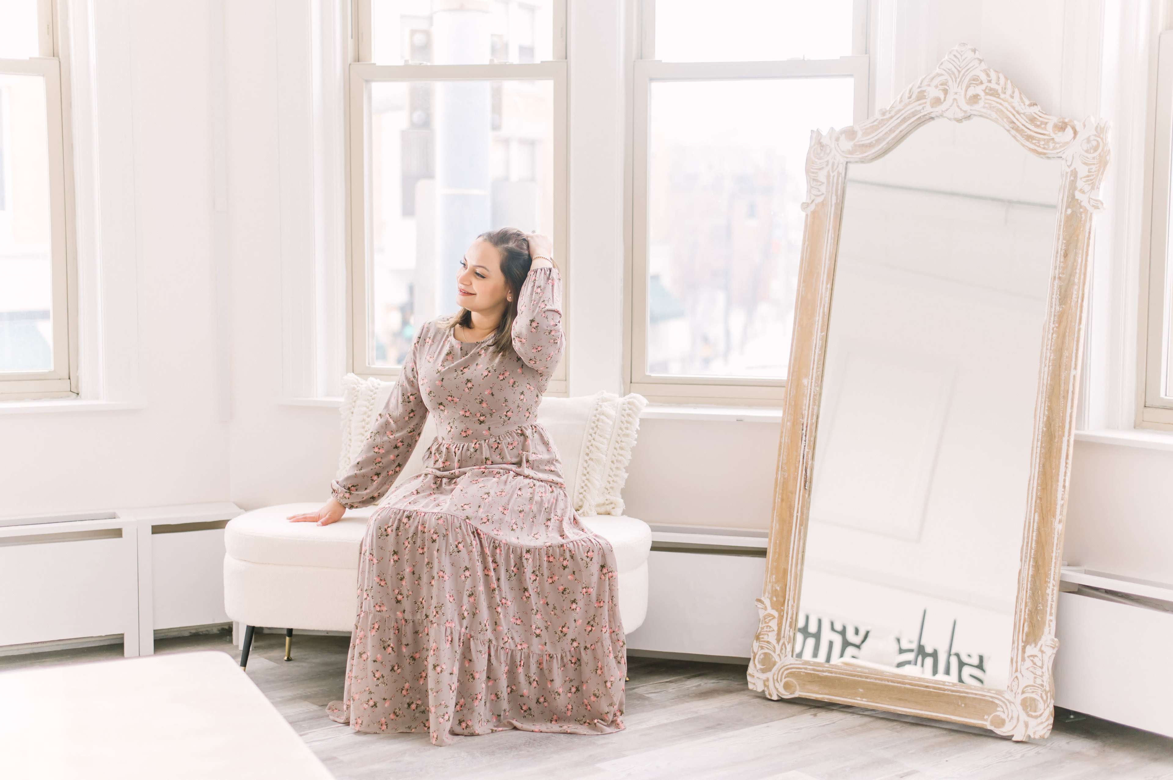 A woman in a long floral dress sits on a cream-colored sofa beside a large, ornate mirror in a bright room with large windows.