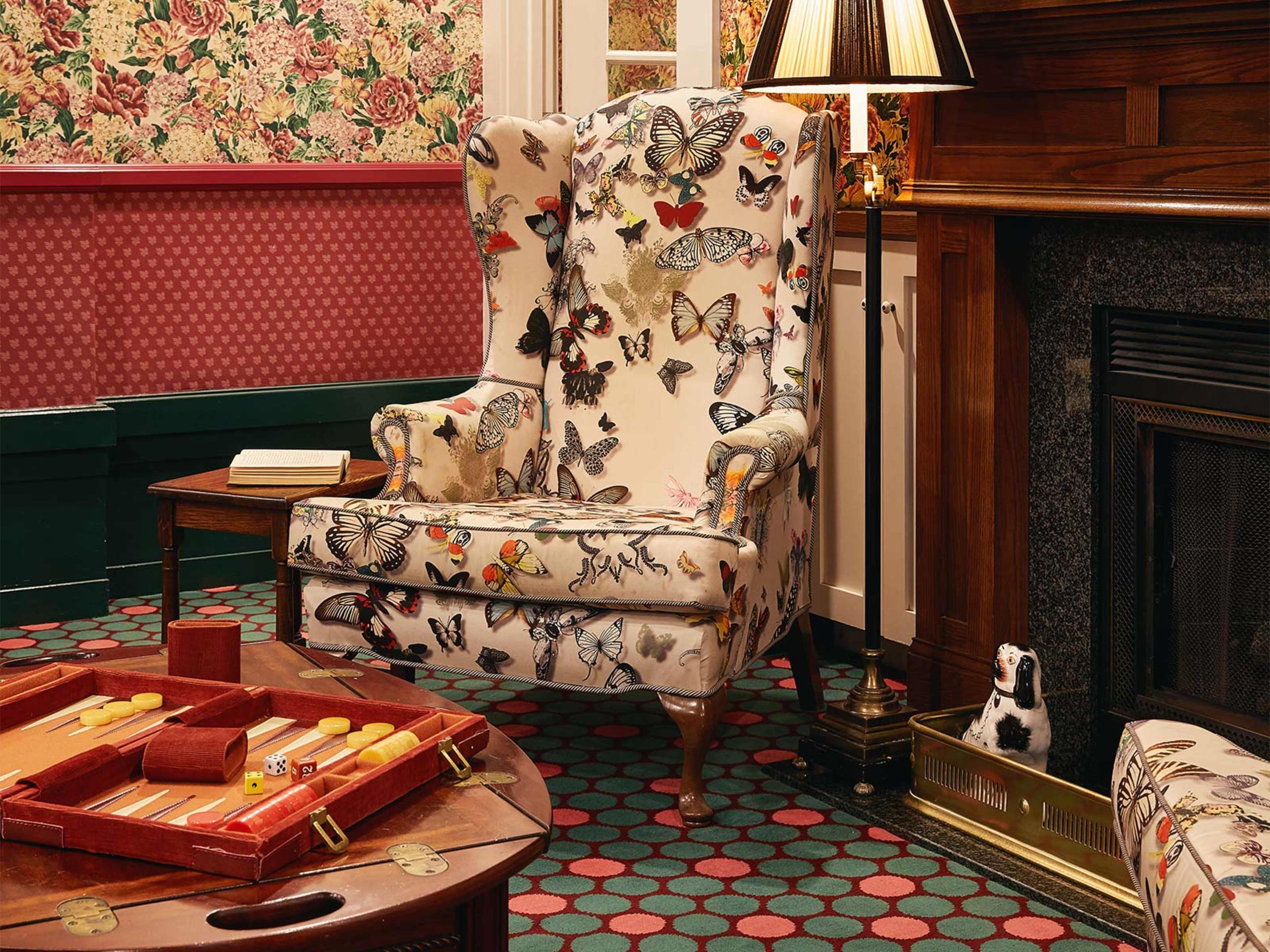 A patterned wingback chair decorated with butterfly motifs, positioned next to a side table and a fireplace in a room with floral wallpaper.