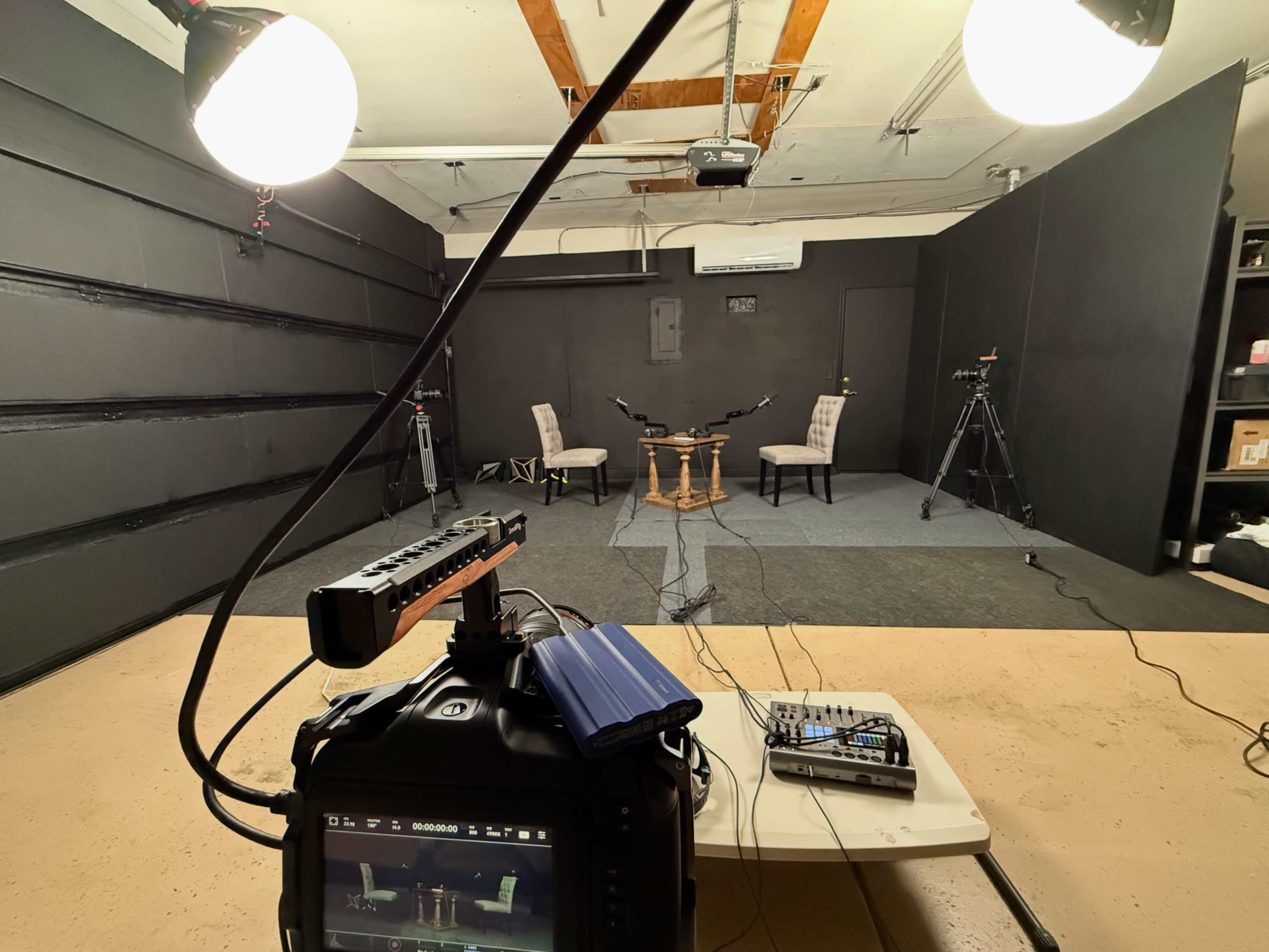 The image shows a studio setup with two chairs and microphones facing each other, surrounded by cameras and lighting equipment on stands.