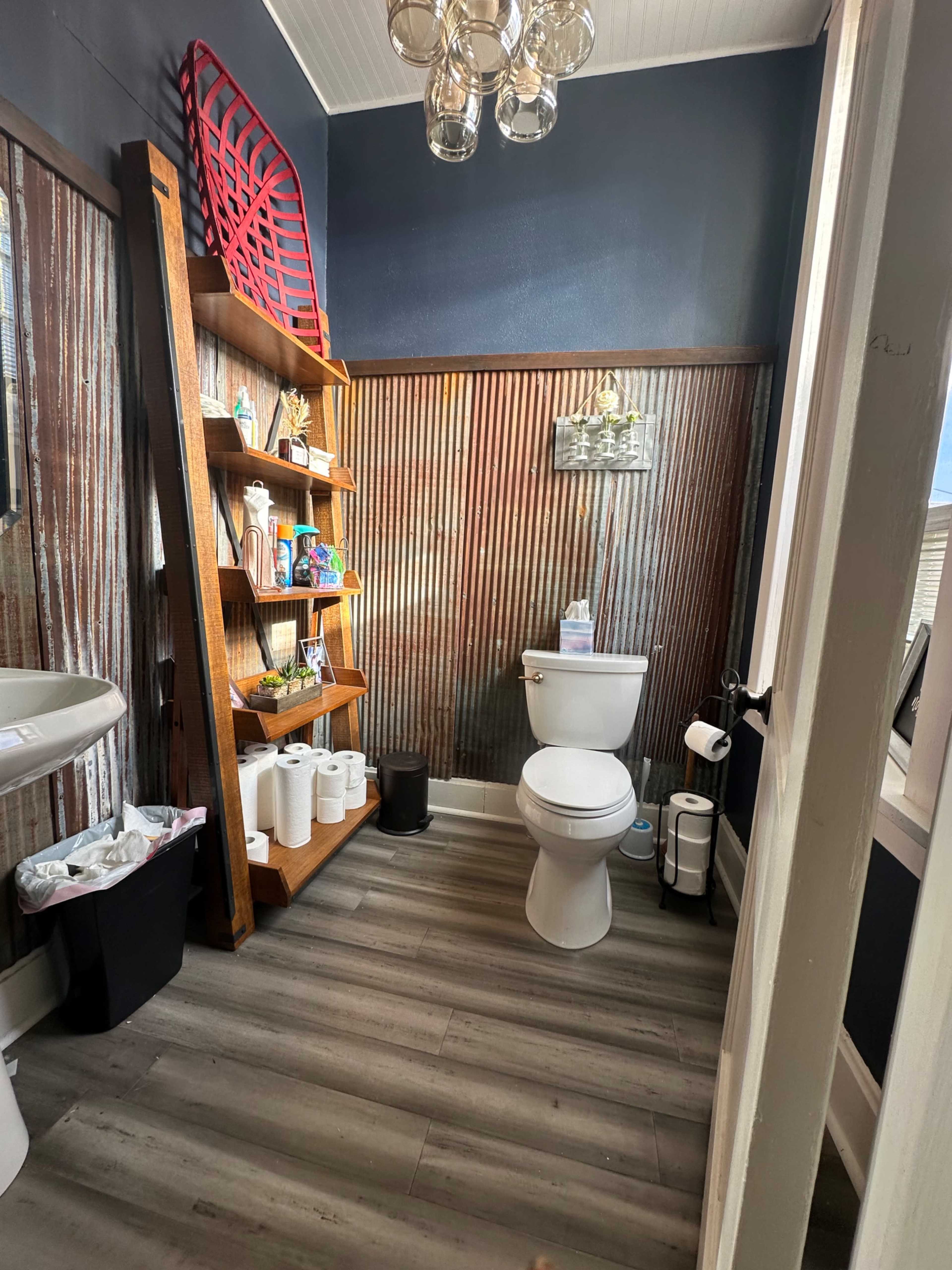 The image shows a compact bathroom with a toilet, a shelf holding various toiletries, and a wall featuring corrugated metal and dark paint.