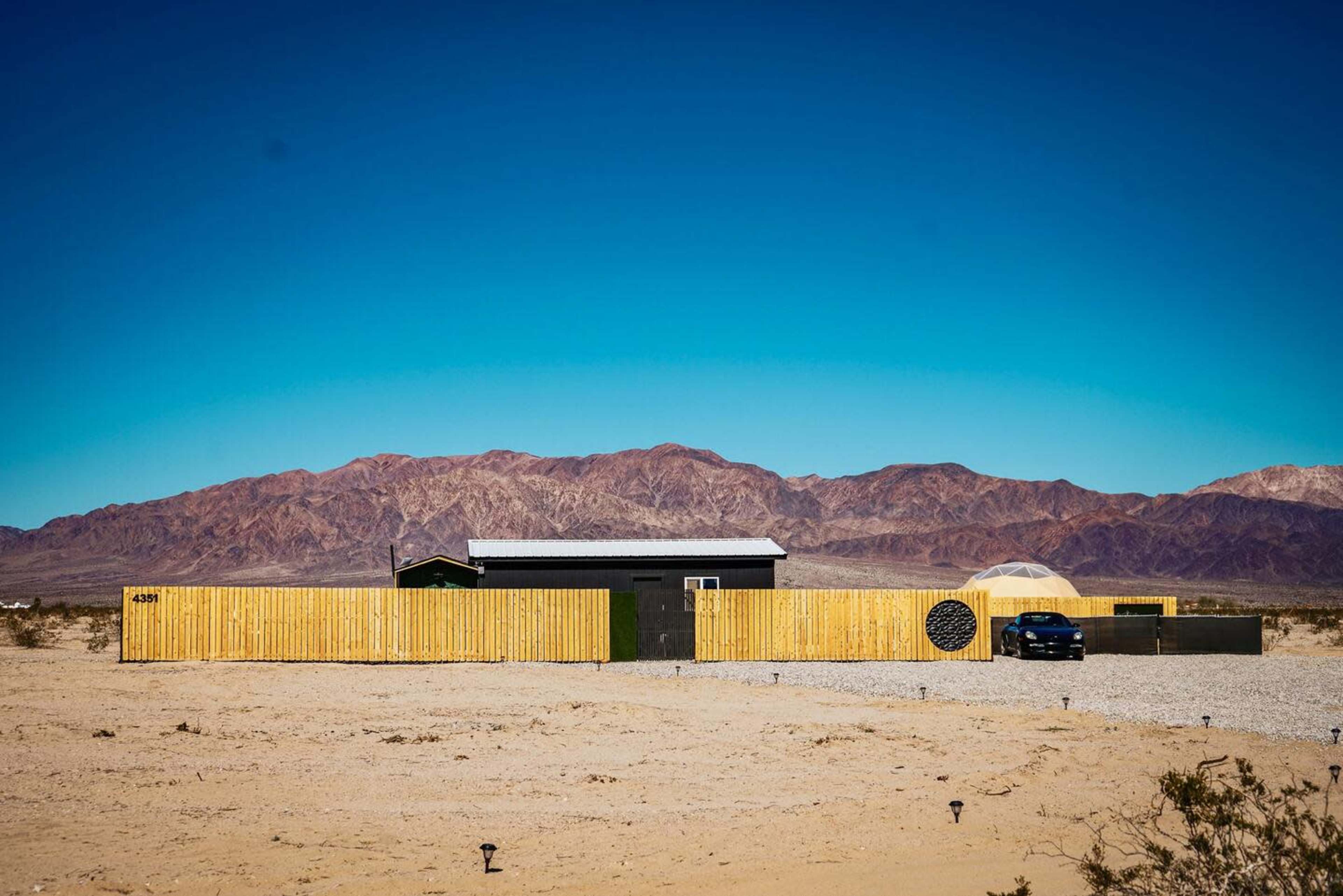 A modern house with a wooden fence is nestled against a backdrop of mountains in a desert landscape.