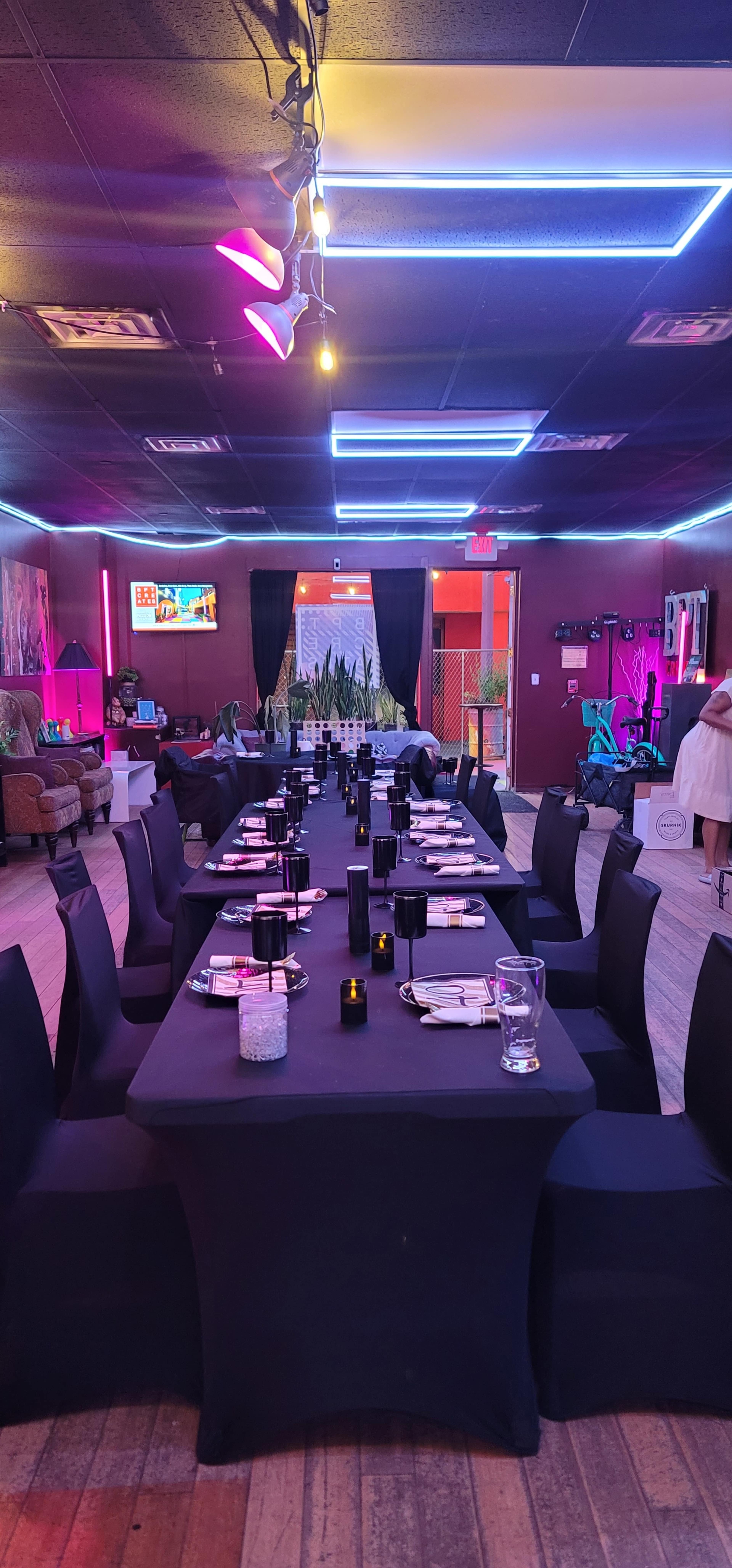 The image shows a dining area set up with black tablecloths, elegant place settings, and ambient lighting, creating a modern atmosphere.