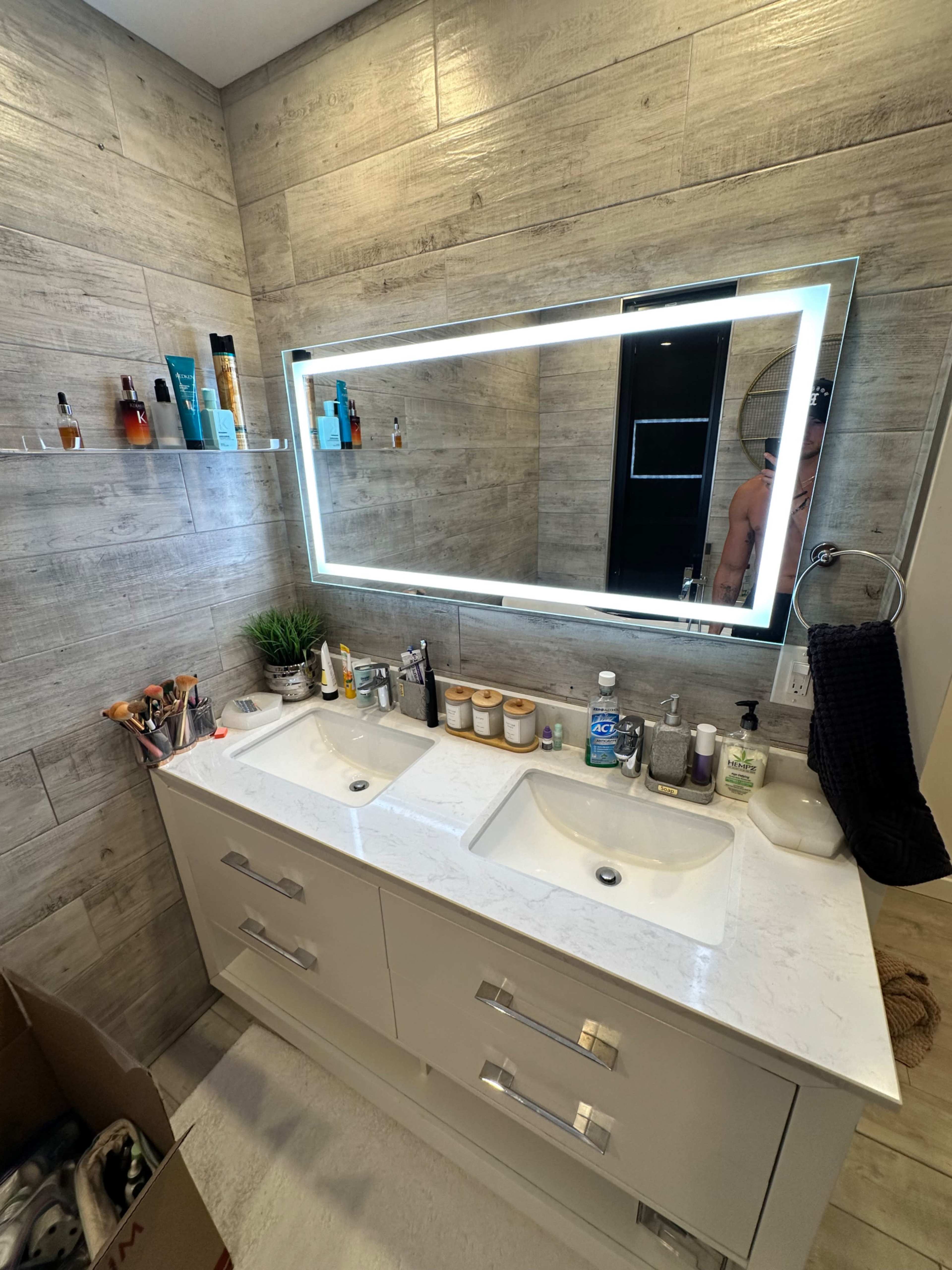 A modern bathroom features a dual sink vanity with a large backlit mirror above, while various toiletries and personal care items are neatly arranged on the countertop and shelves.