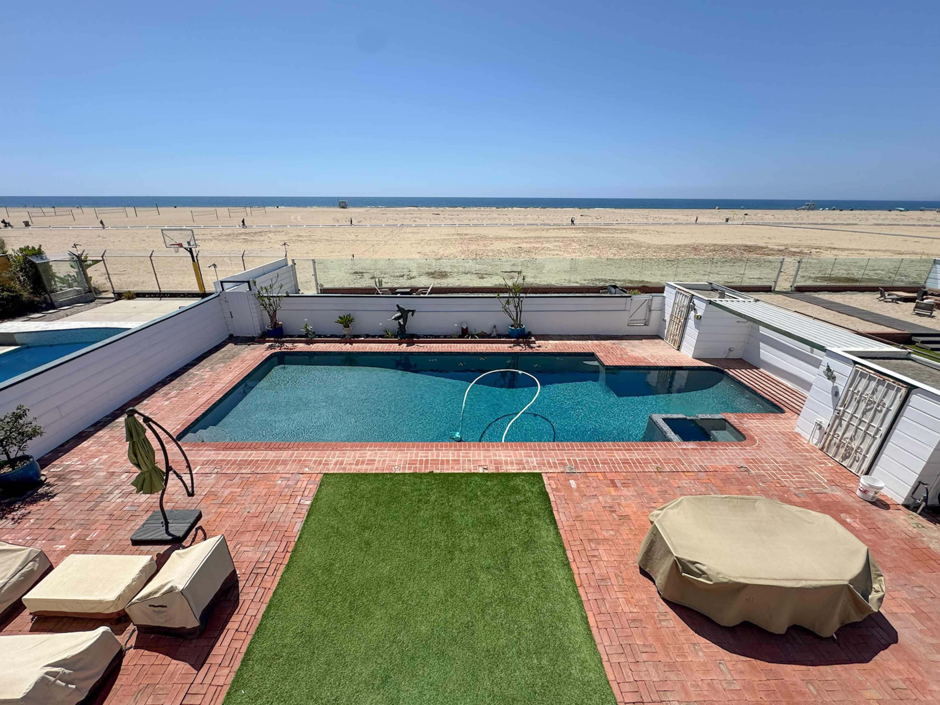 The image features a beachfront property with a swimming pool, surrounded by lounge chairs and manicured grass, overlooking the sandy beach and ocean.