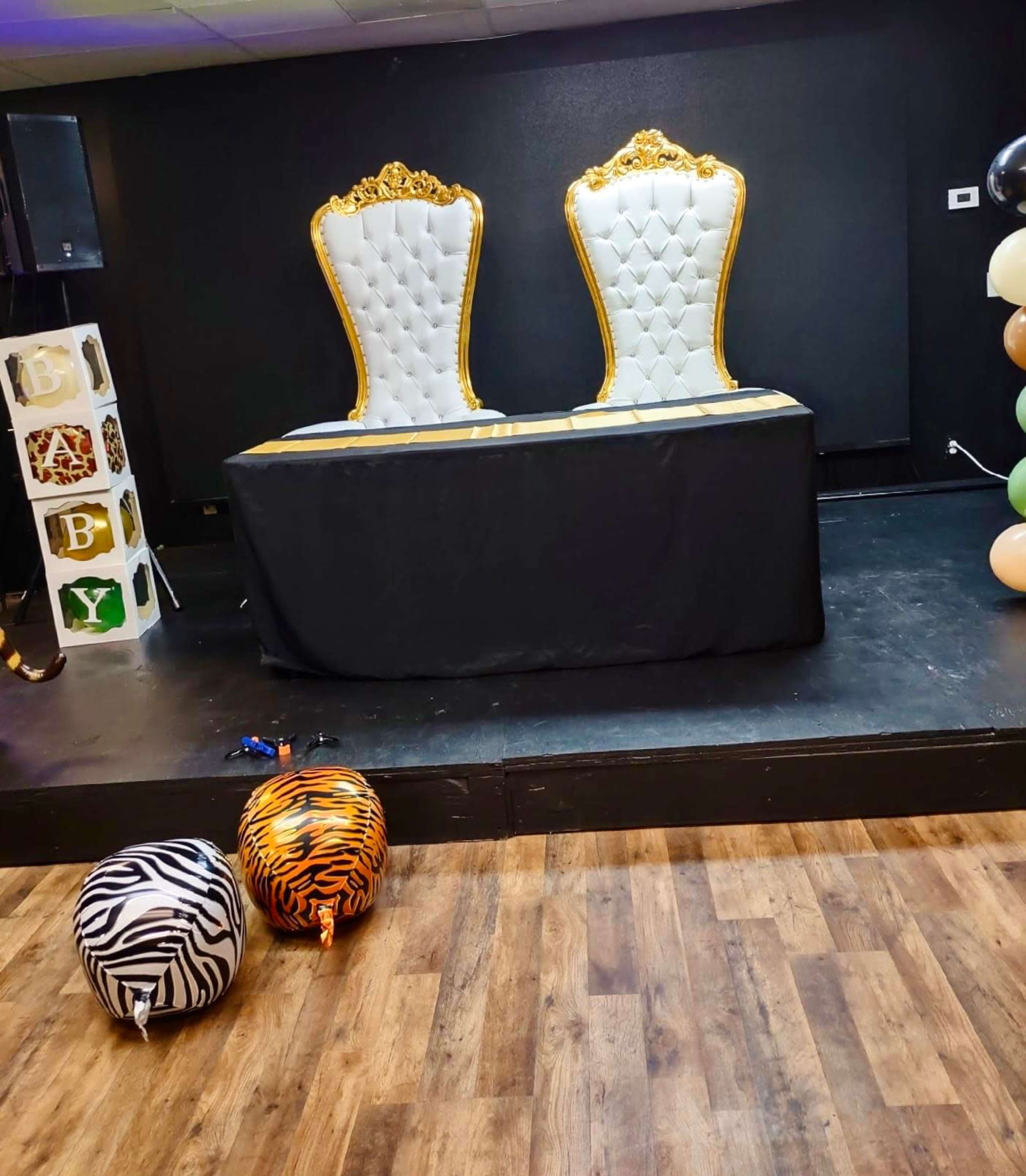 A decorated event space featuring two elegant white chairs with gold frames on a stage, accompanied by a black table and two patterned poufs in front.