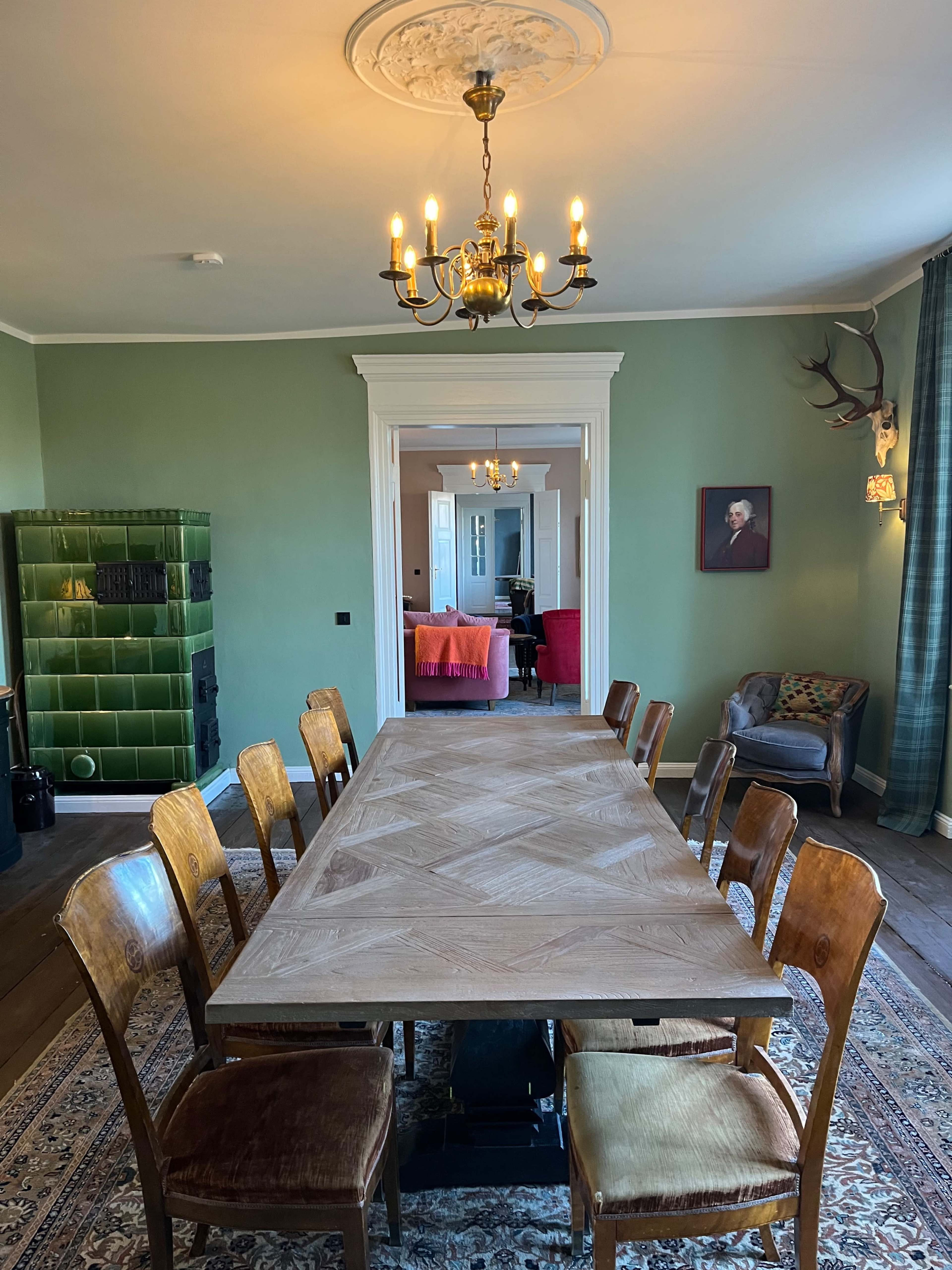 The image shows a spacious dining room with a large wooden table surrounded by chairs, featuring green walls, a chandelier, and a doorway leading to another room with colorful furnishings.