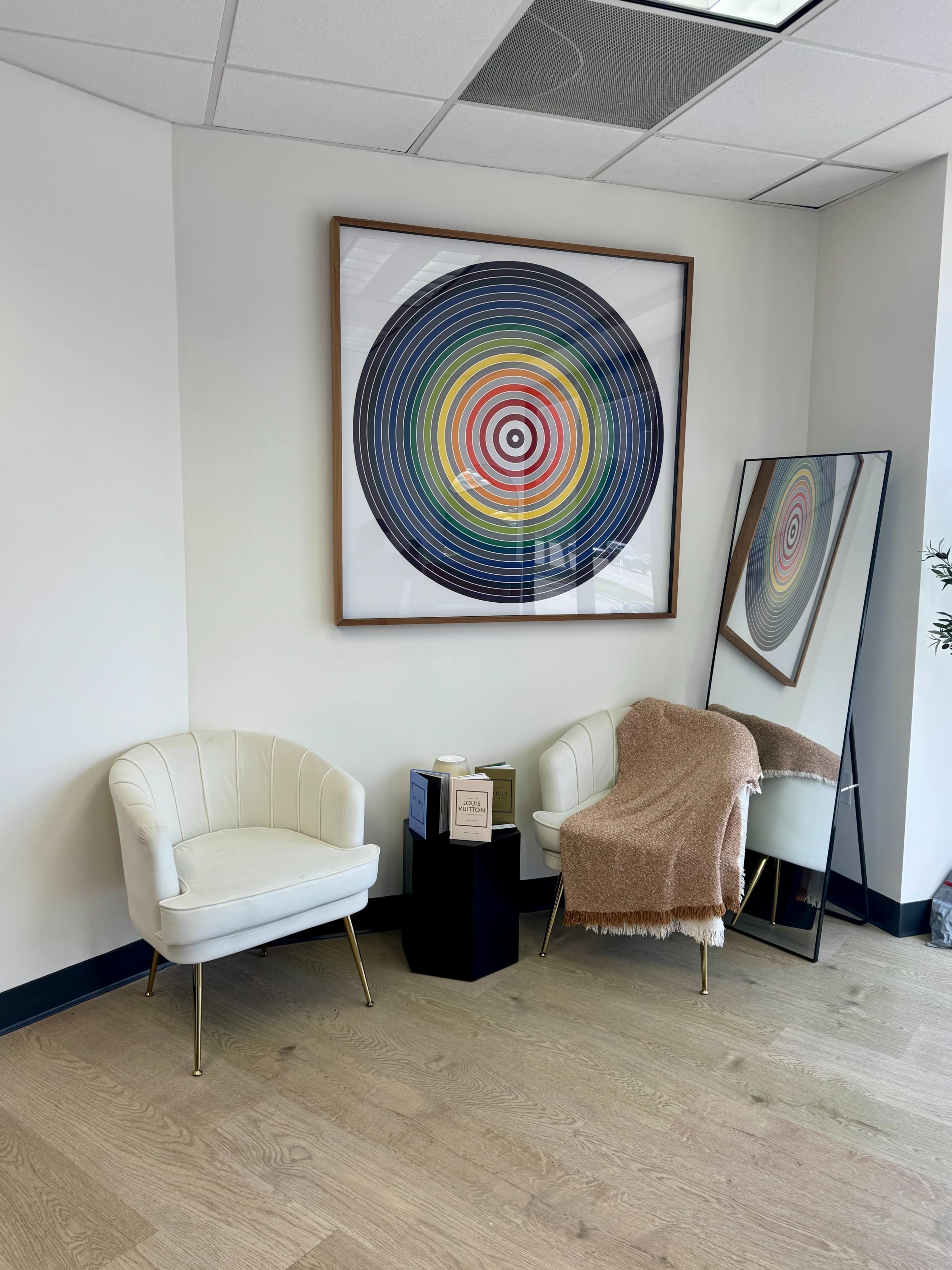 The image shows a corner of an office featuring two white chairs, a small side table, a colorful circular artwork on the wall, and a large mirror.