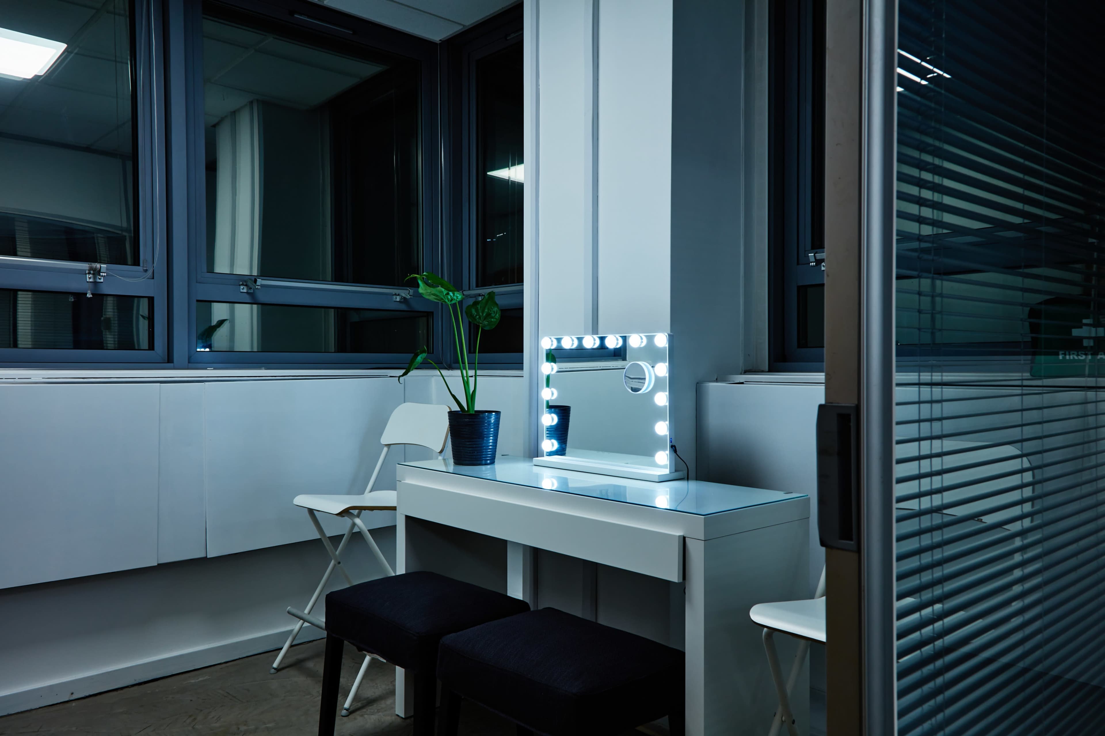 A bright vanity mirror with lights is positioned on a table, accompanied by two black stools and a potted plant, in a room with large windows.