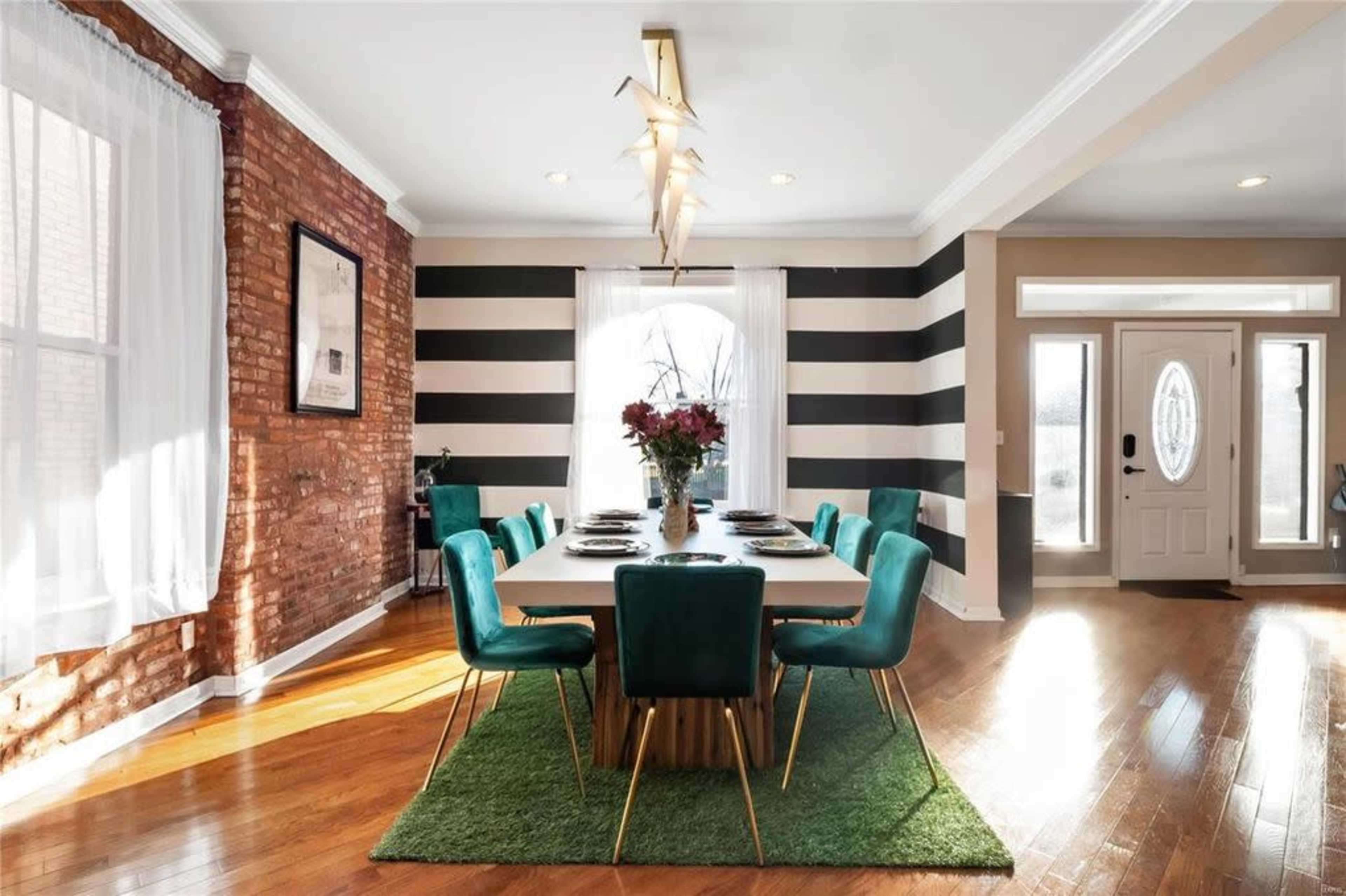 A modern dining room features a long table surrounded by green chairs, positioned on a green rug, with striped walls and large windows allowing natural light.