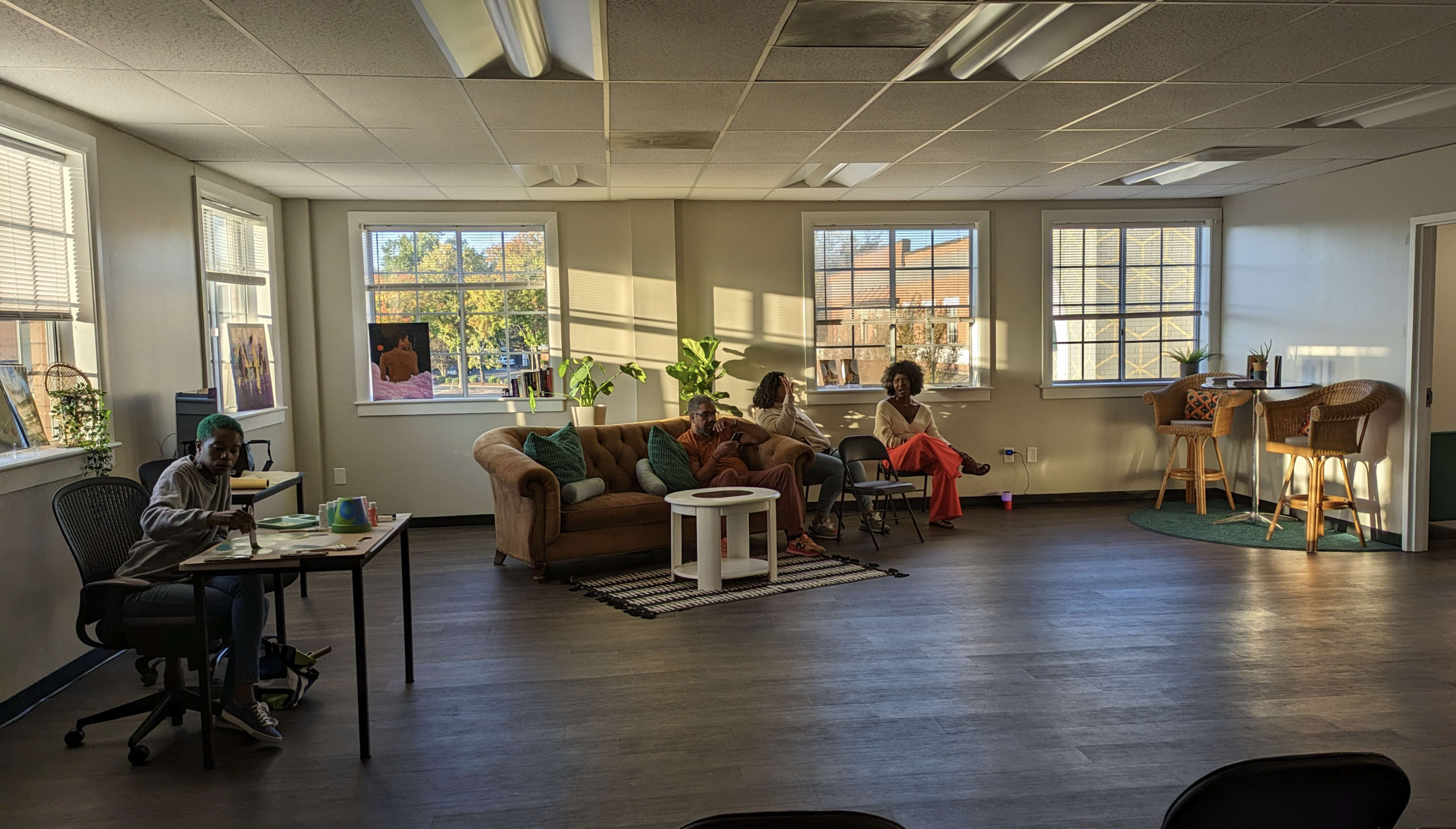 A spacious, well-lit room features a couch, chairs, tables, and large windows, with three people seated in different areas.