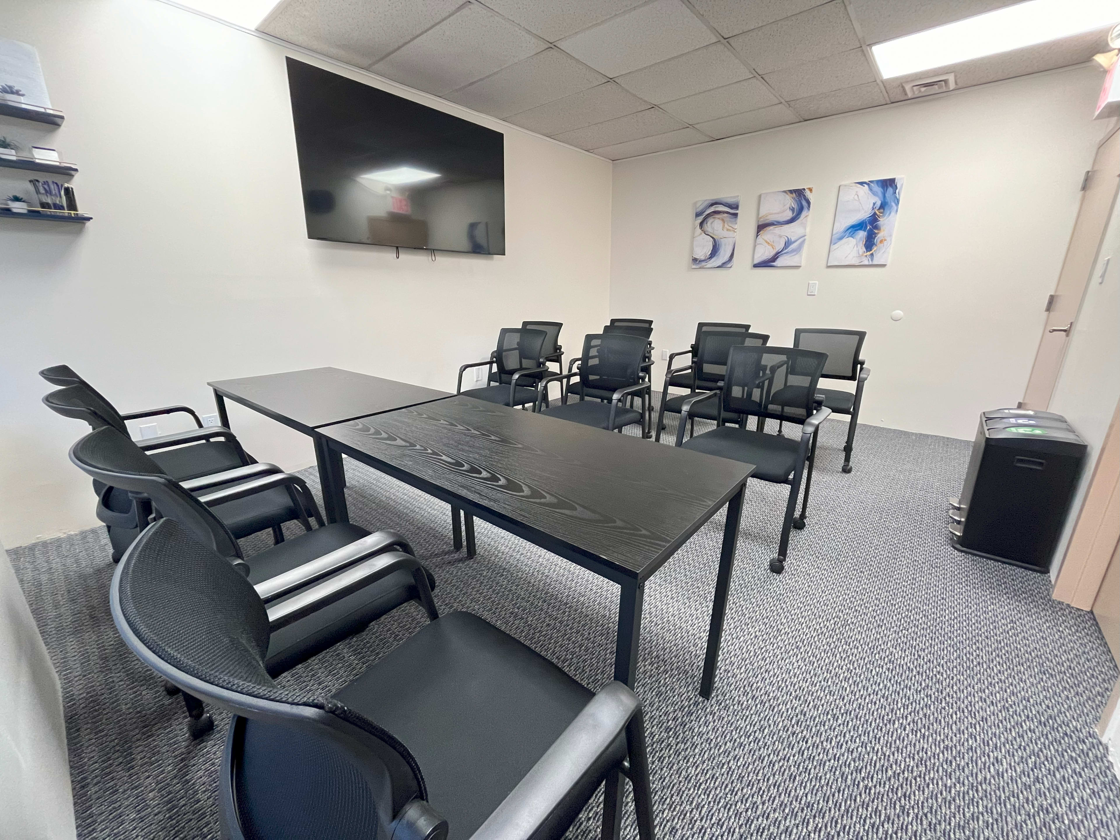 A conference room features a long black table surrounded by multiple black chairs, with a large screen mounted on the wall and abstract artwork displayed nearby.