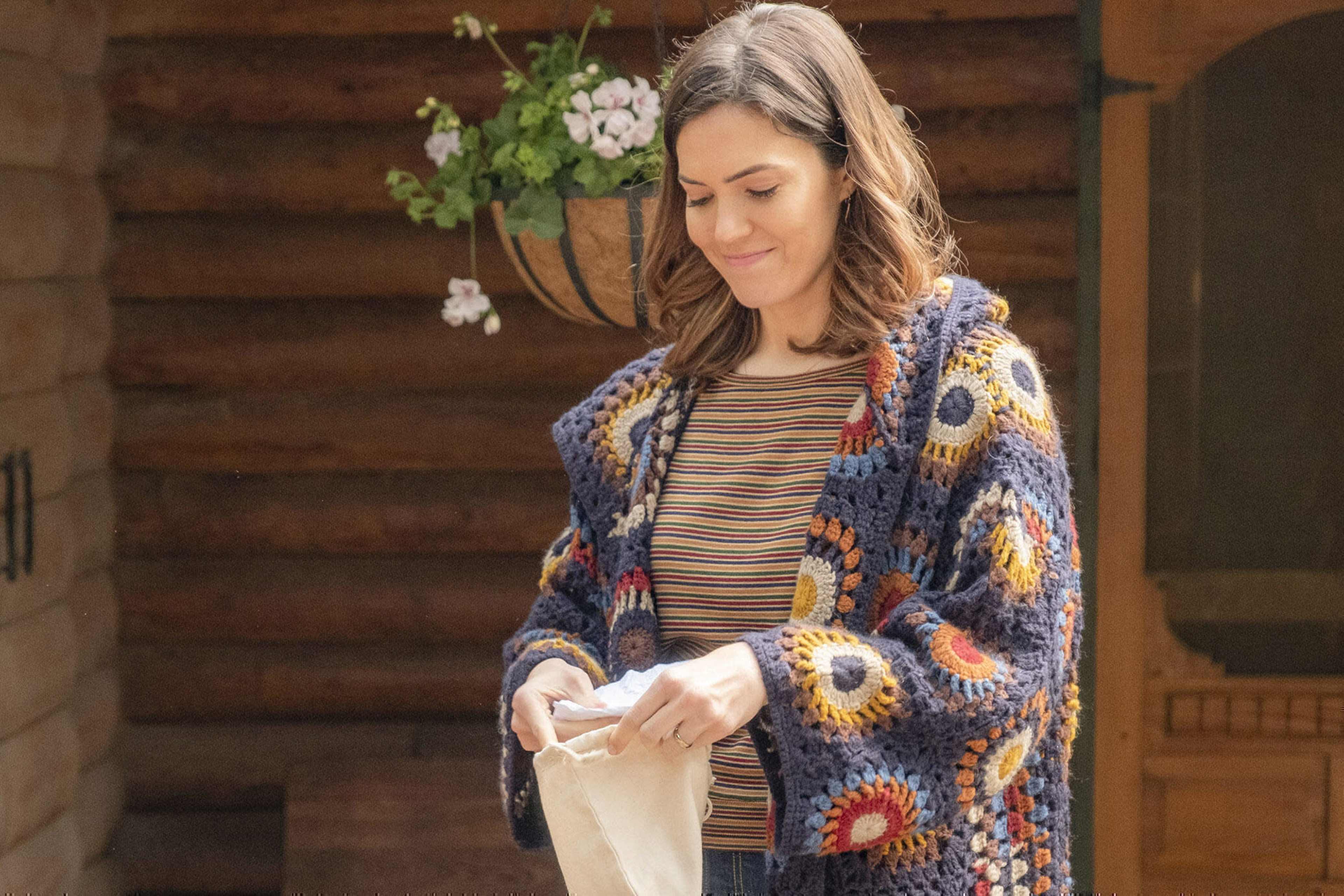 A woman in a colorful, knitted cardigan smiles while holding a small cloth bag in front of a wooden cabin.