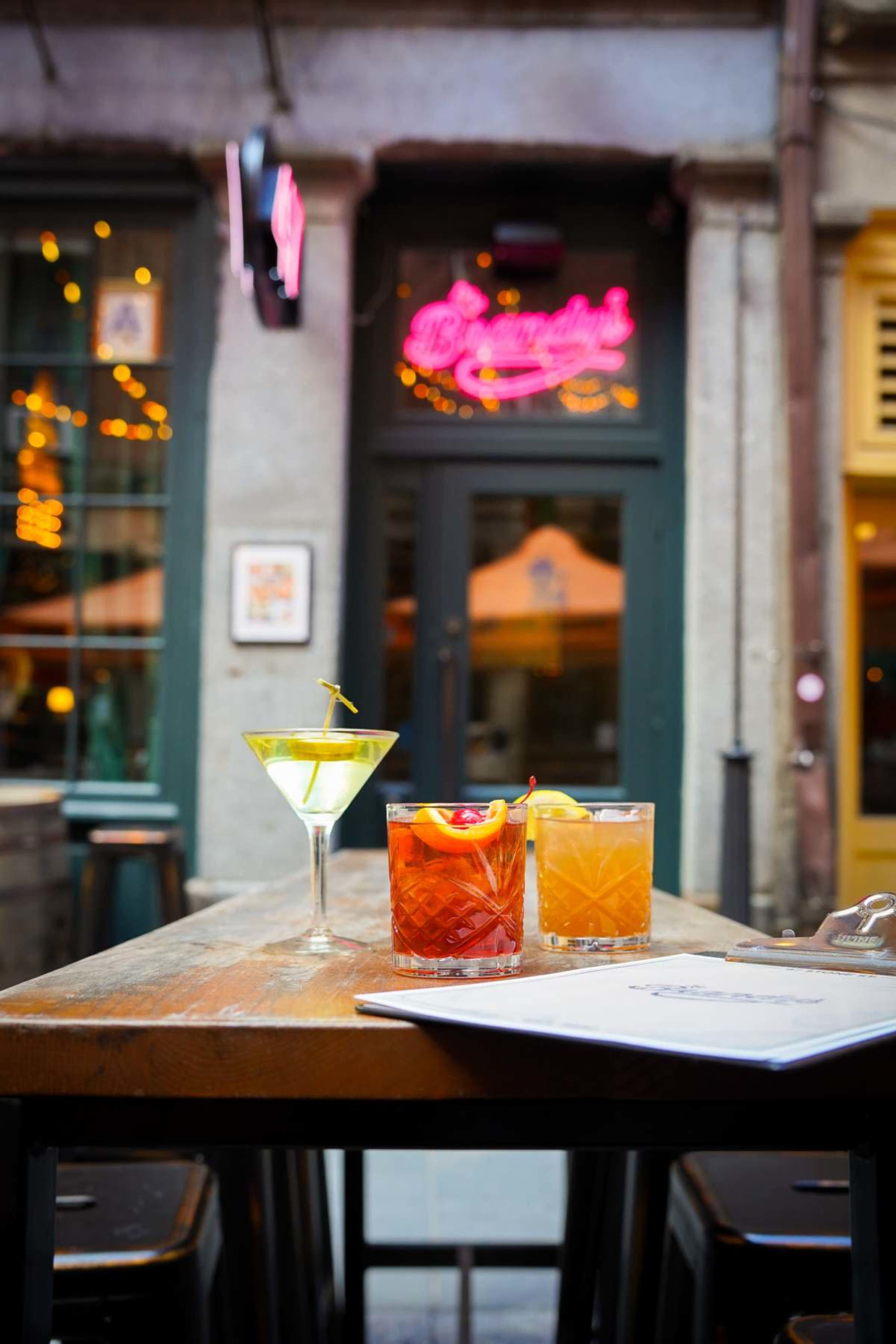 Three cocktails are placed on a wooden table outside a bar, with a neon sign in the background.