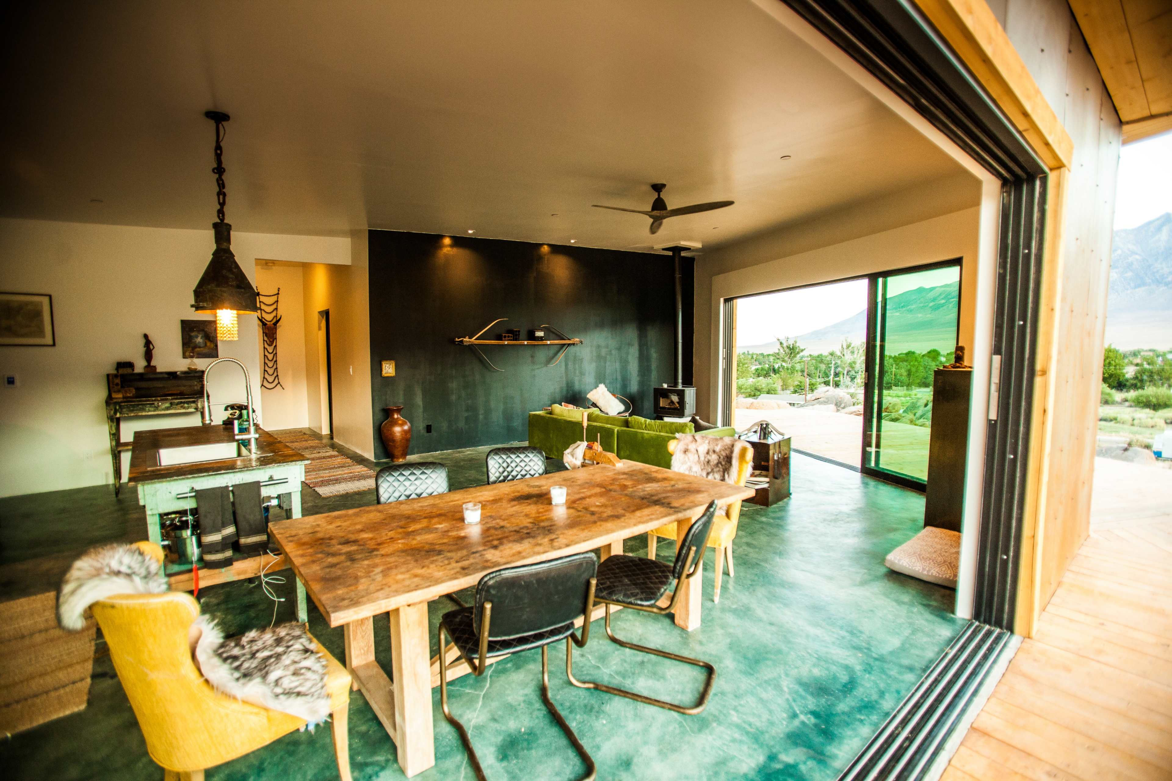 The image shows a modern dining area with a wooden table surrounded by chairs, large sliding doors opening to a scenic outdoor landscape, and minimalist furnishings.