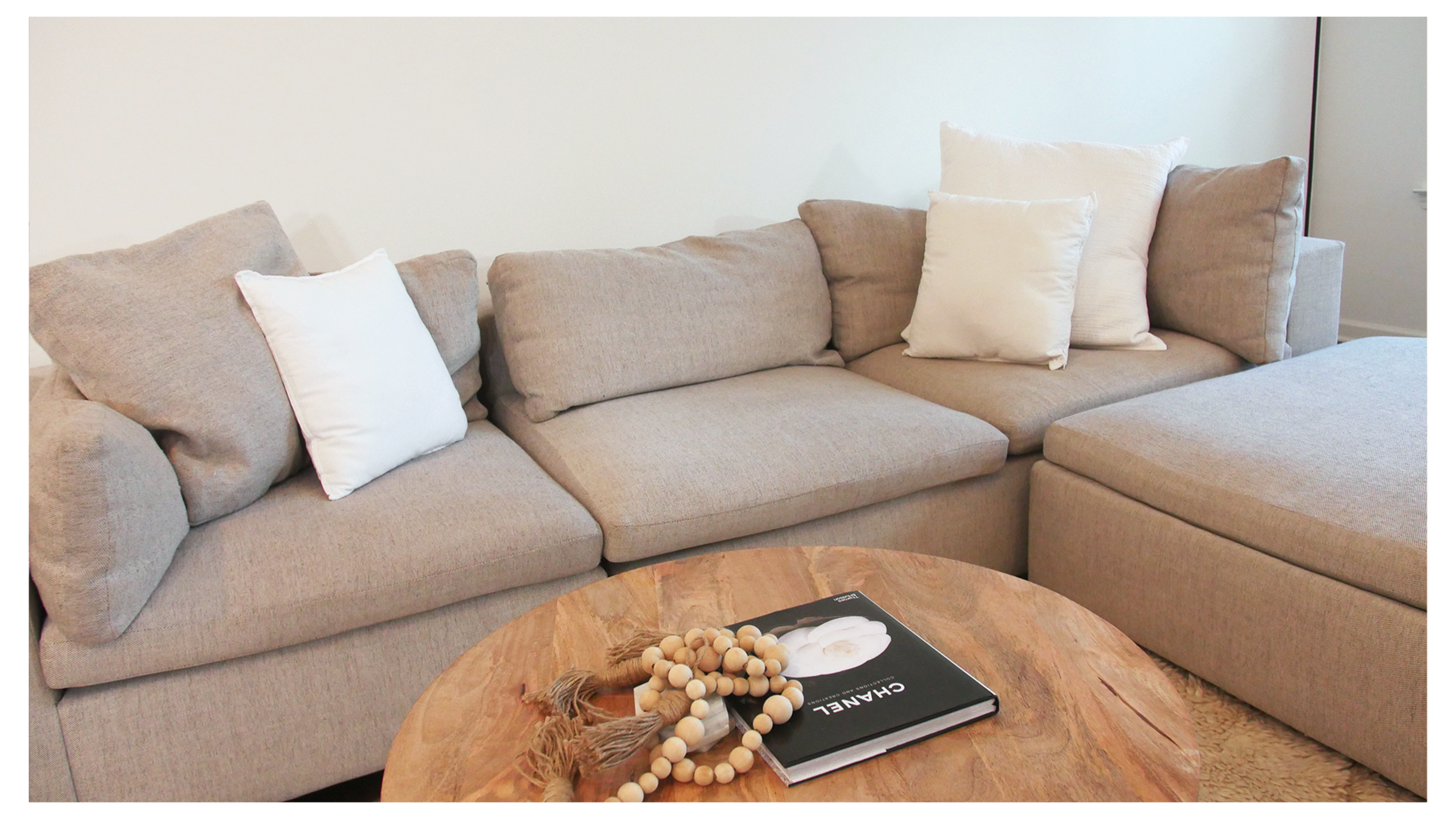 A light gray sectional sofa with white pillows is positioned beside a wooden coffee table featuring a book and decorative beads.