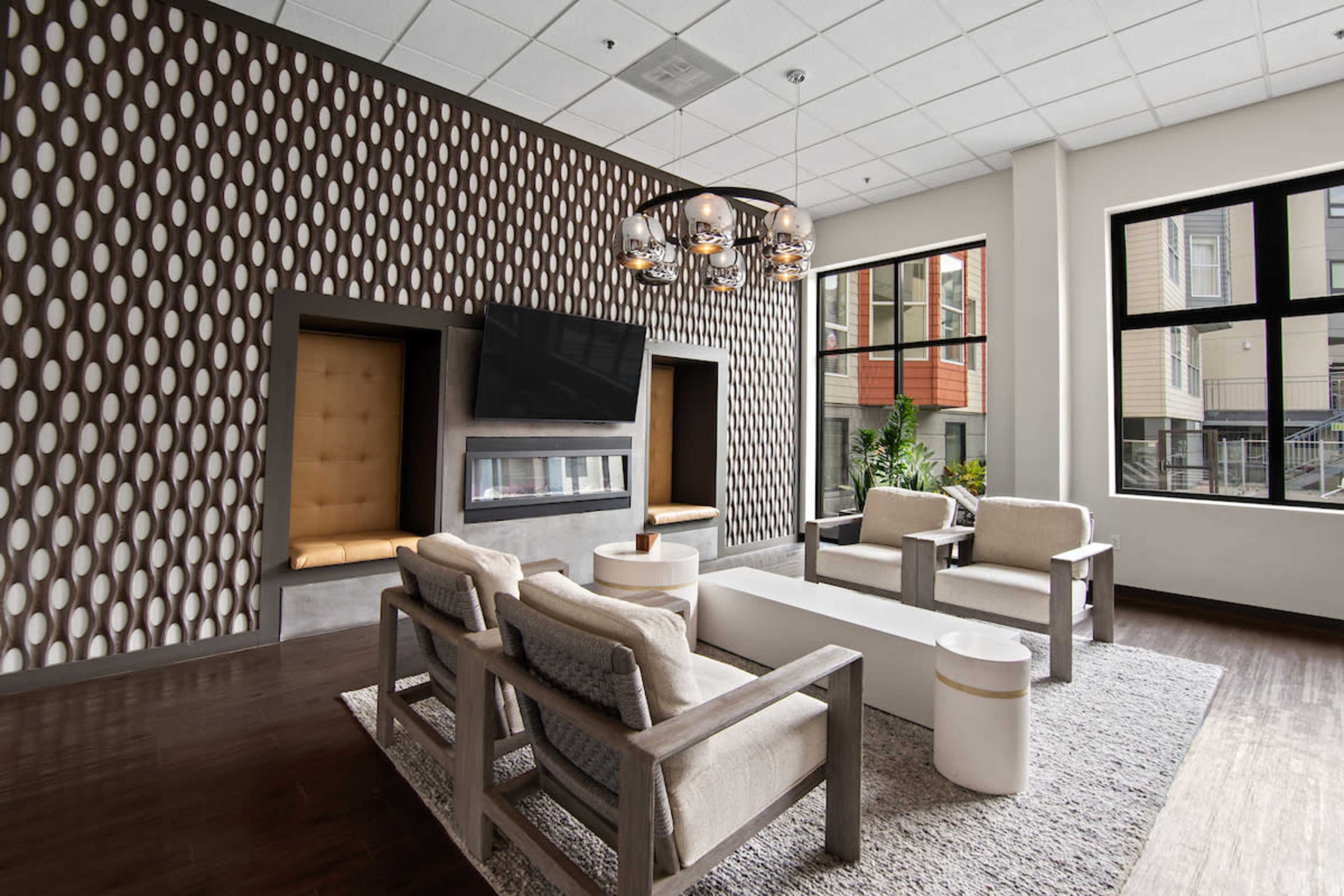 The image shows a modern lounge area featuring two upholstered seating spaces, a central white coffee table, and a large television mounted above a fireplace, all surrounded by large windows.