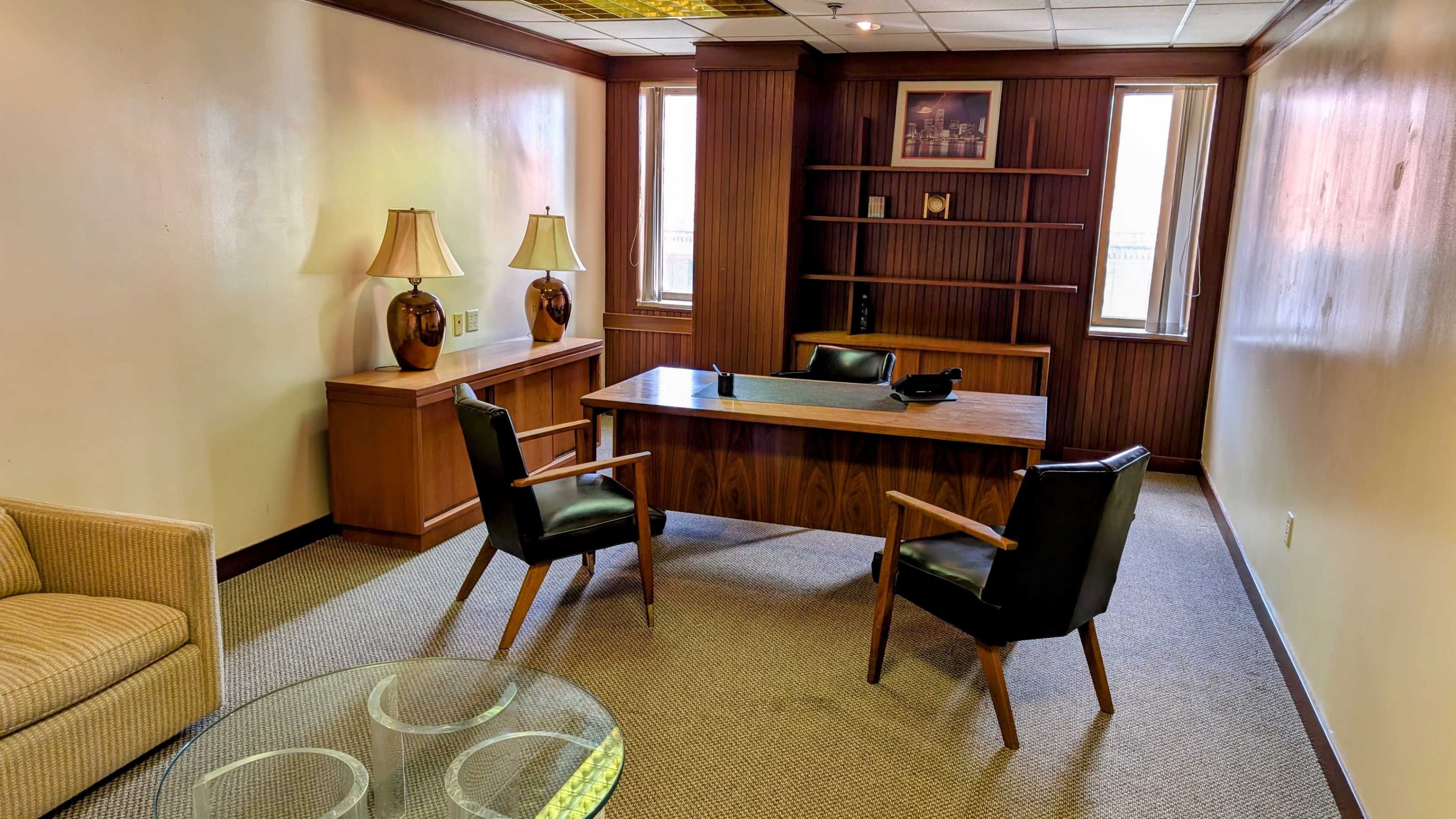 The image shows a small office with a wooden desk, two black chairs, a glass coffee table, and two lamps, set against light-colored walls and featuring built-in shelves.