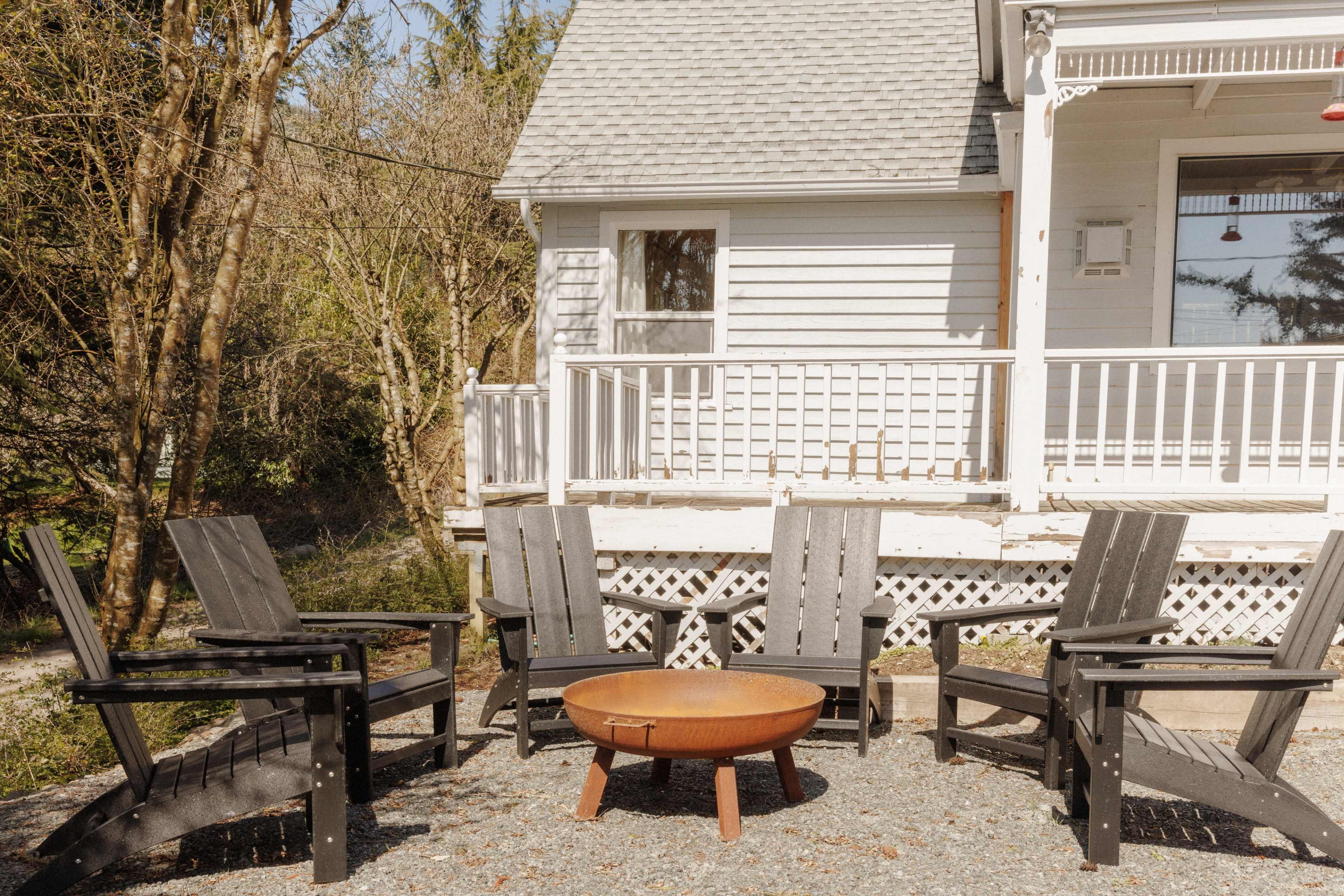 A set of four black Adirondack chairs surrounds a circular fire pit on a gravel area next to a house with a white exterior and a porch.