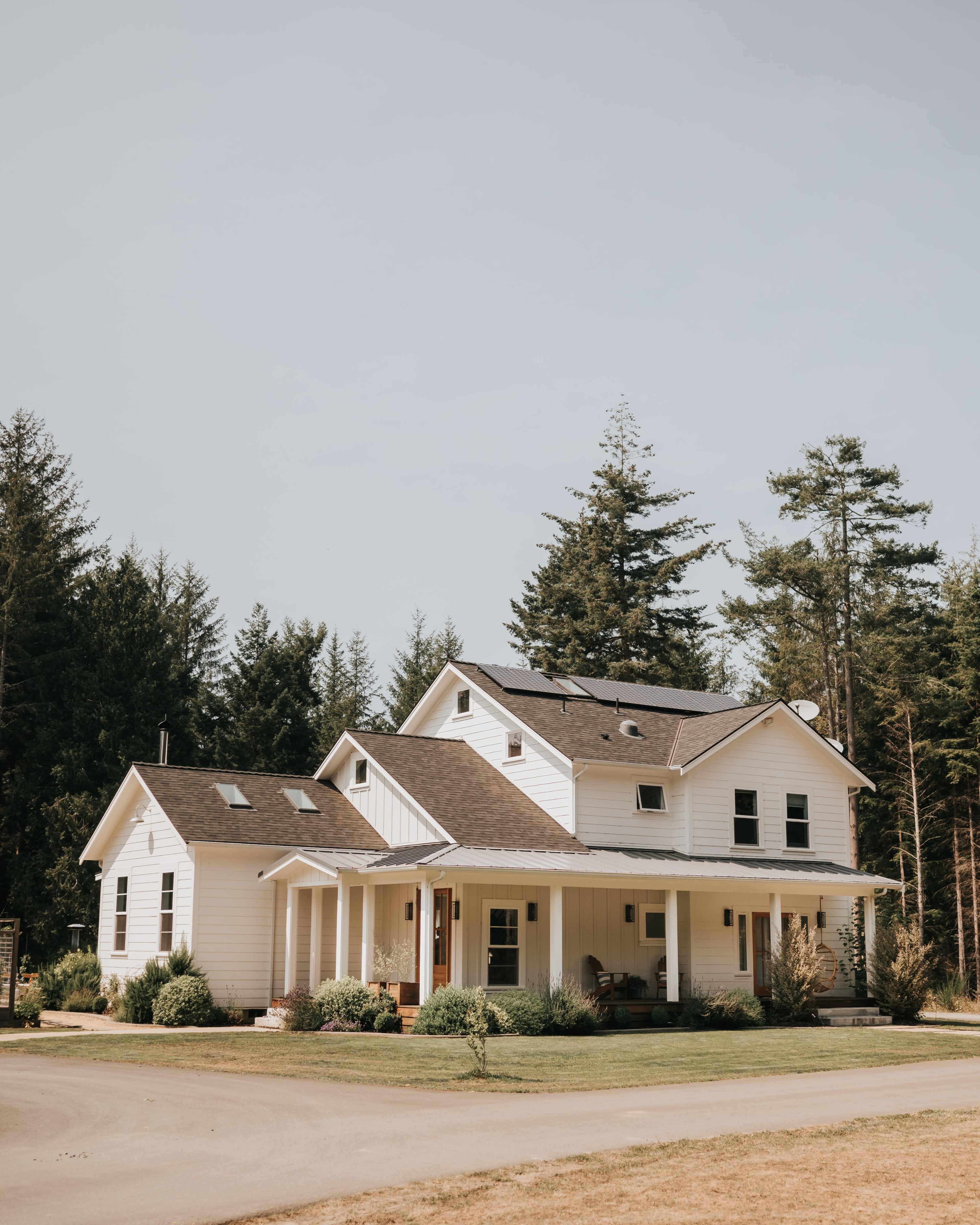 A white, two-story house with a sloped roof is surrounded by trees and features a porch and solar panels on the roof.