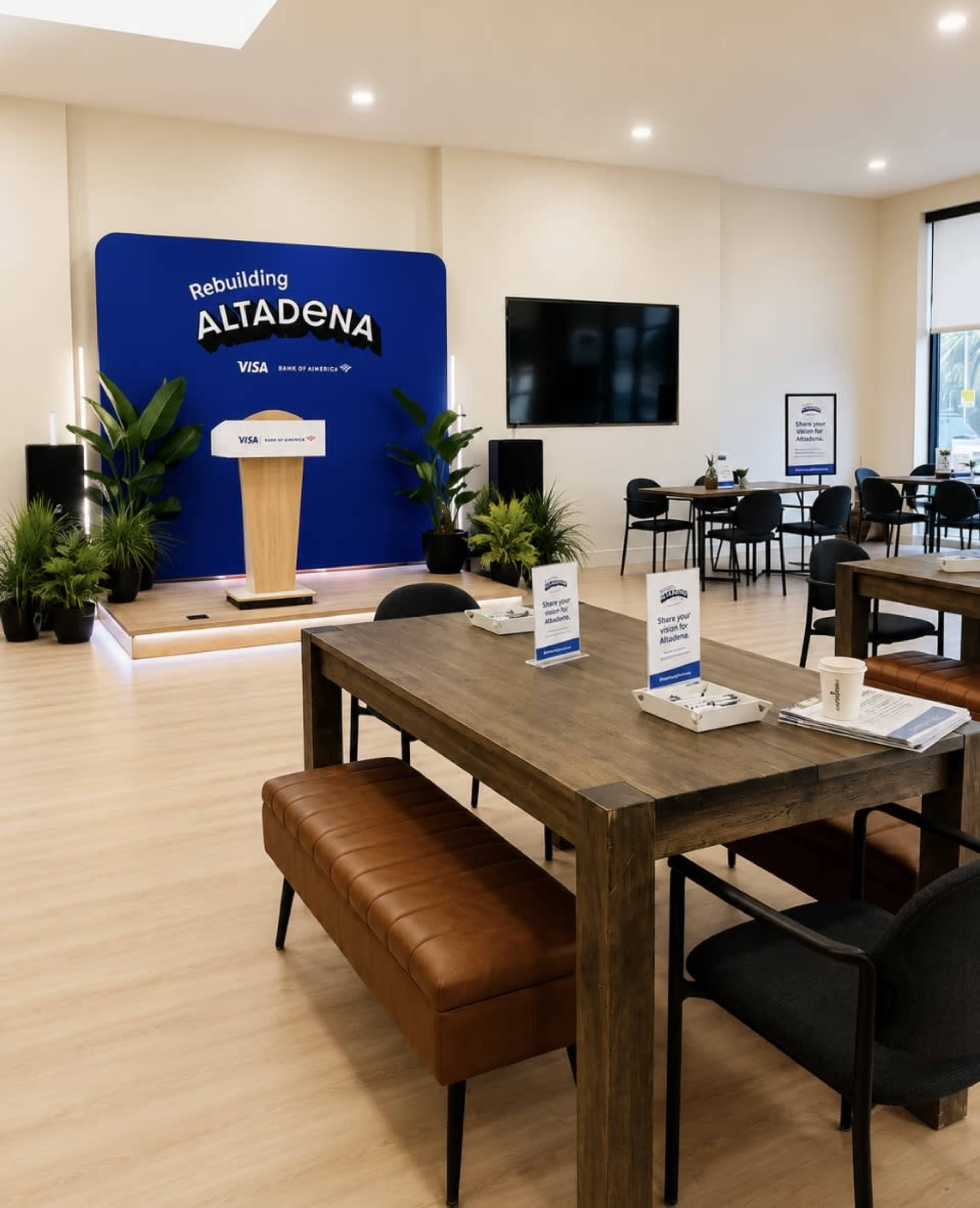The image shows a modern event space featuring a podium with a backdrop that reads "Rebuilding Altadena," surrounded by tables and chairs.
