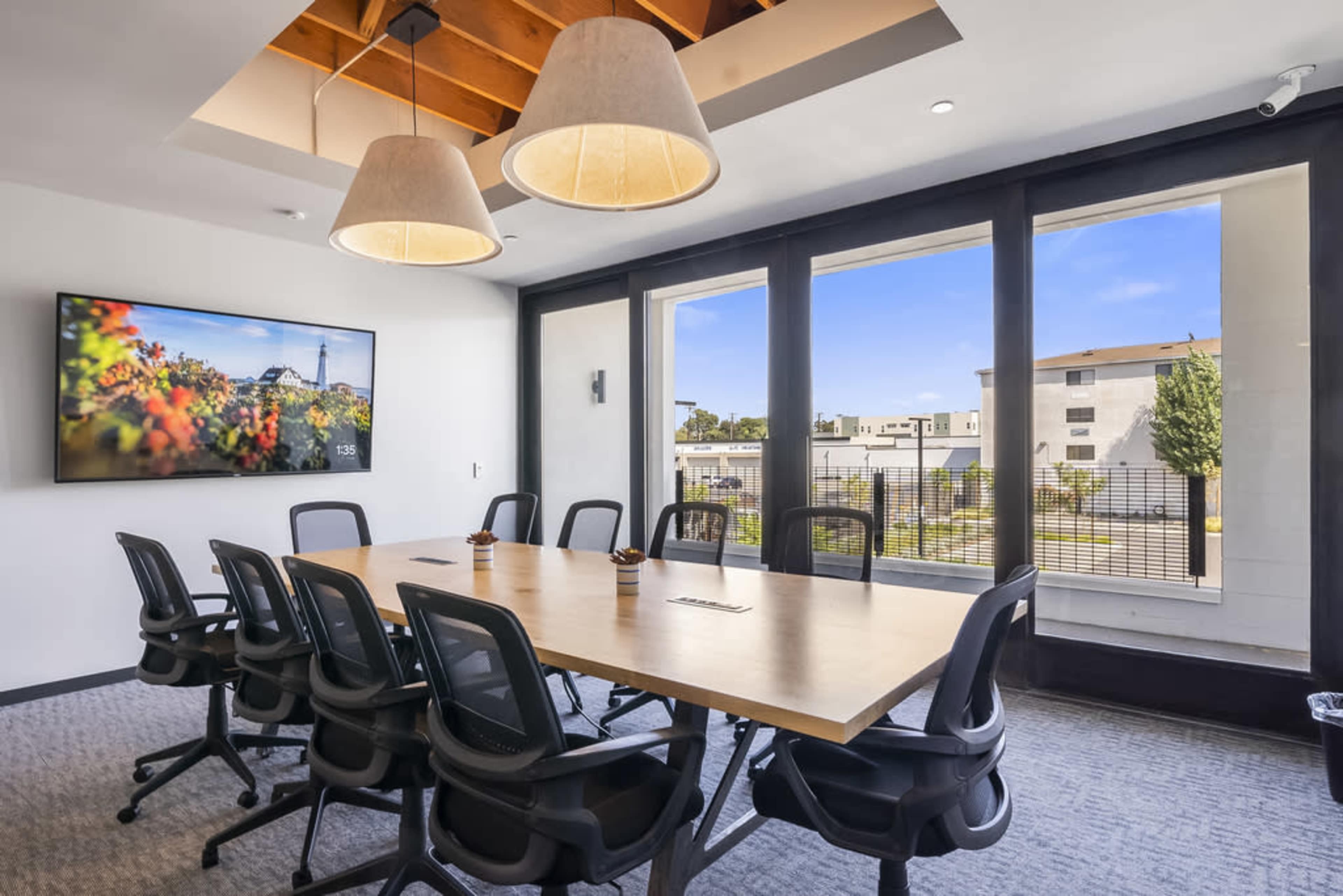 A conference room features a long wooden table surrounded by ergonomic chairs, with a large screen on one wall and windows providing natural light.