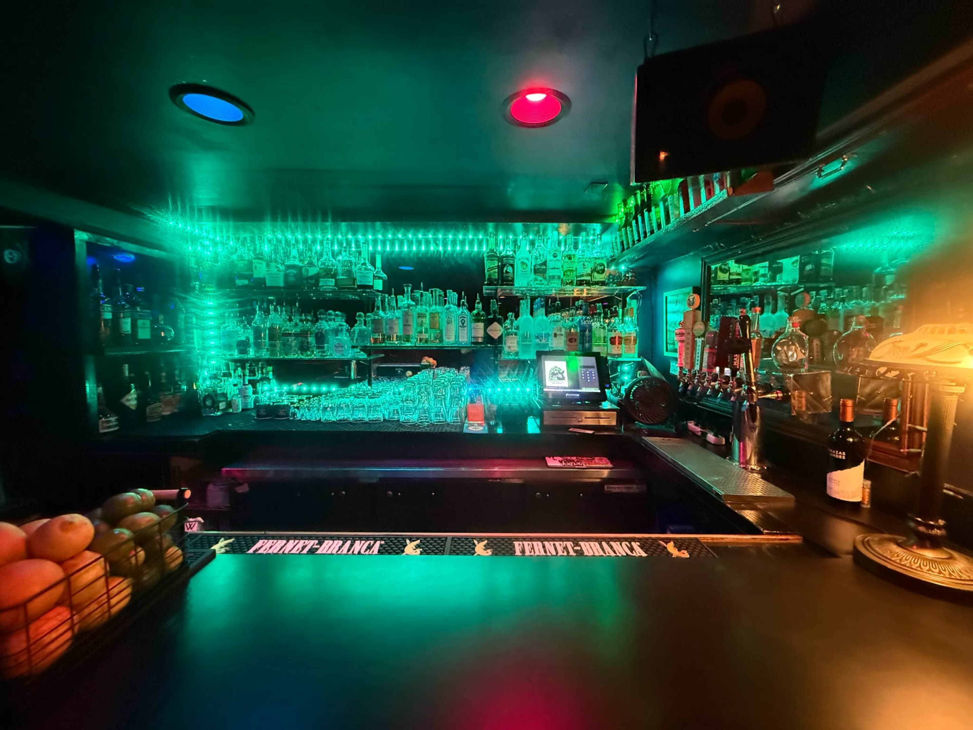 The image shows a dimly lit bar with shelves stocked full of colorful bottles behind the counter, illuminated by green and red LED lights.