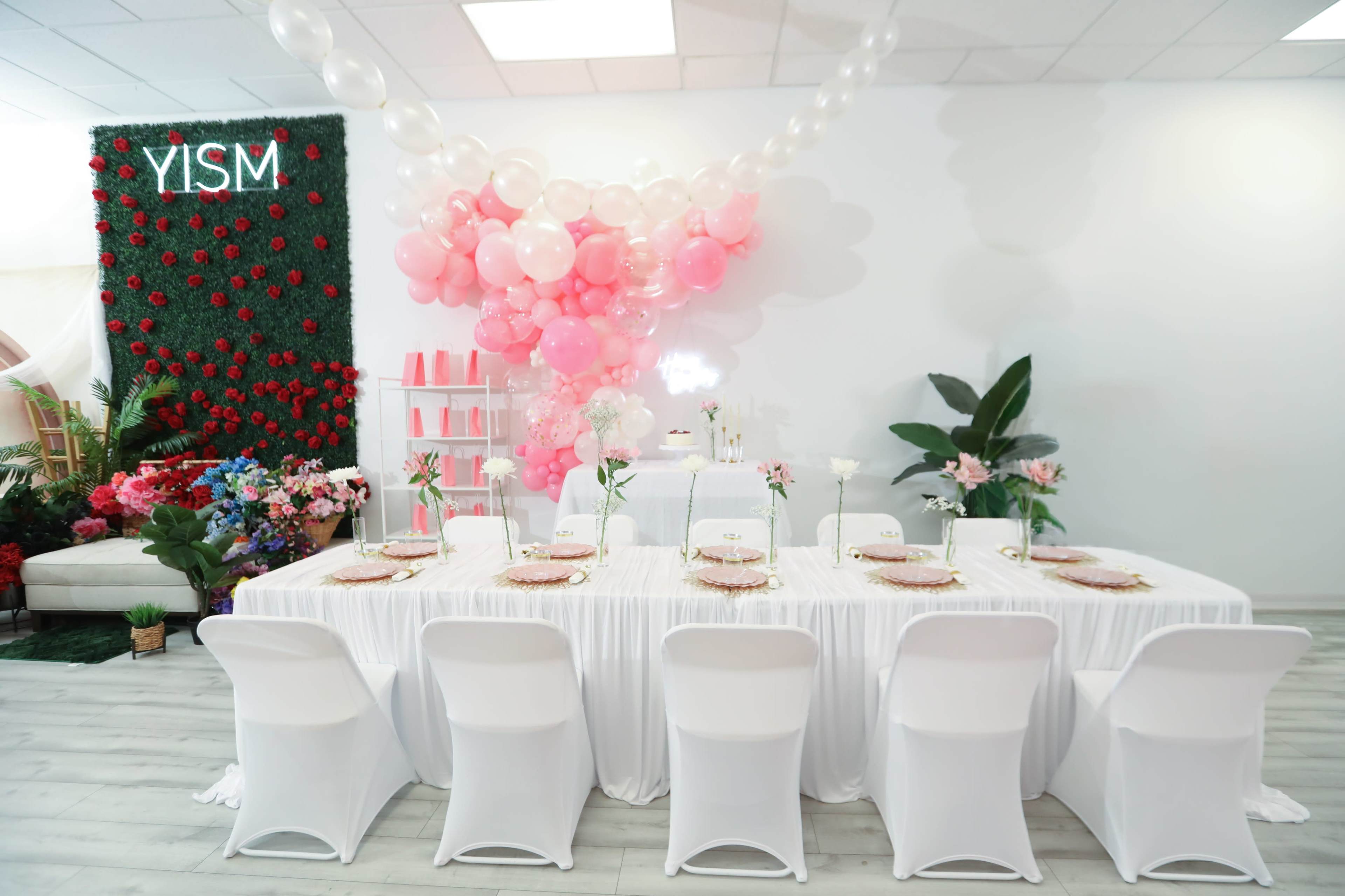 A long table with white linens is set for a celebration, adorned with pink balloons and floral arrangements, against a backdrop of greenery and decorative elements.