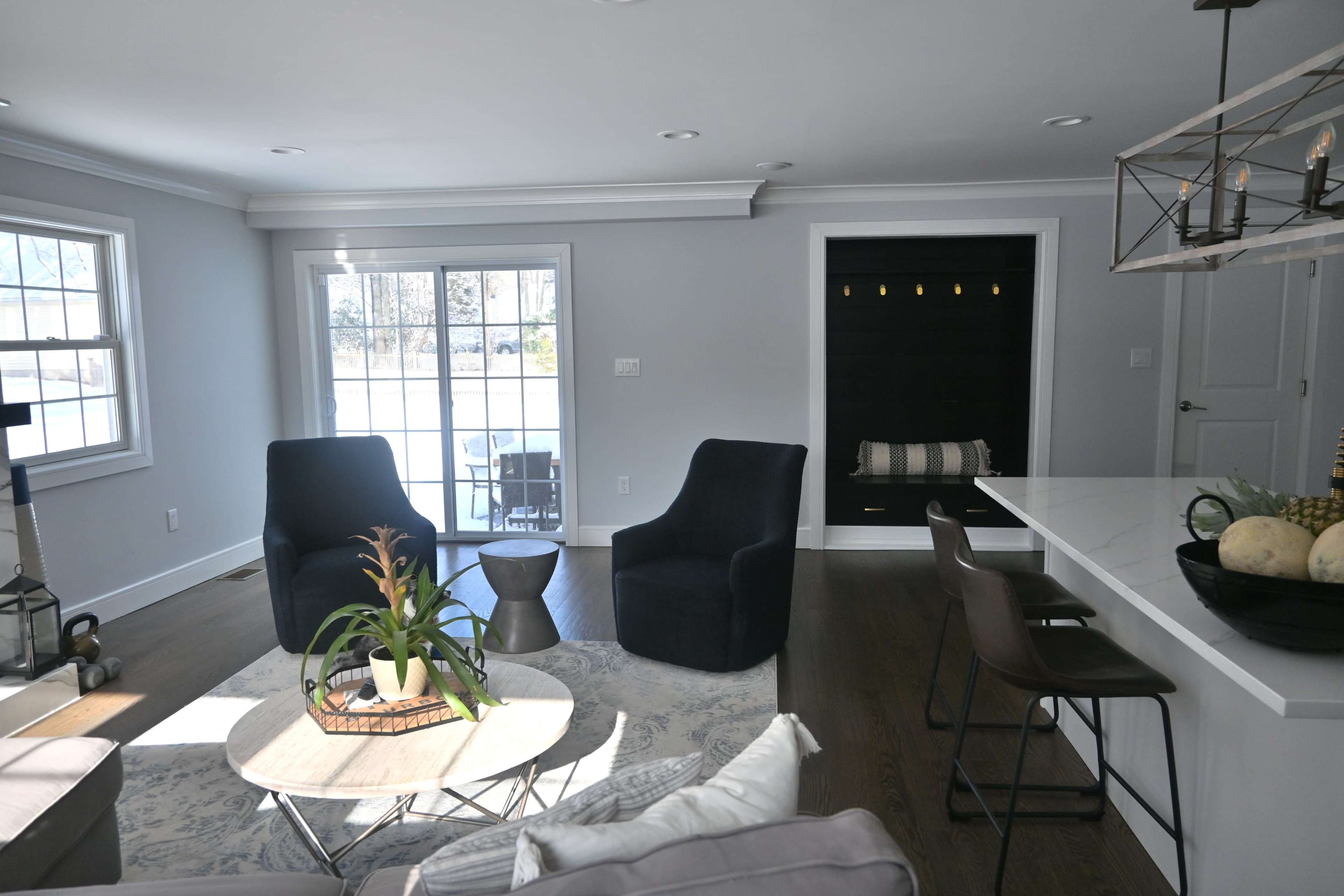 A modern living room with two dark chairs facing a coffee table, a large window letting in natural light, and a kitchen bar area with stools.