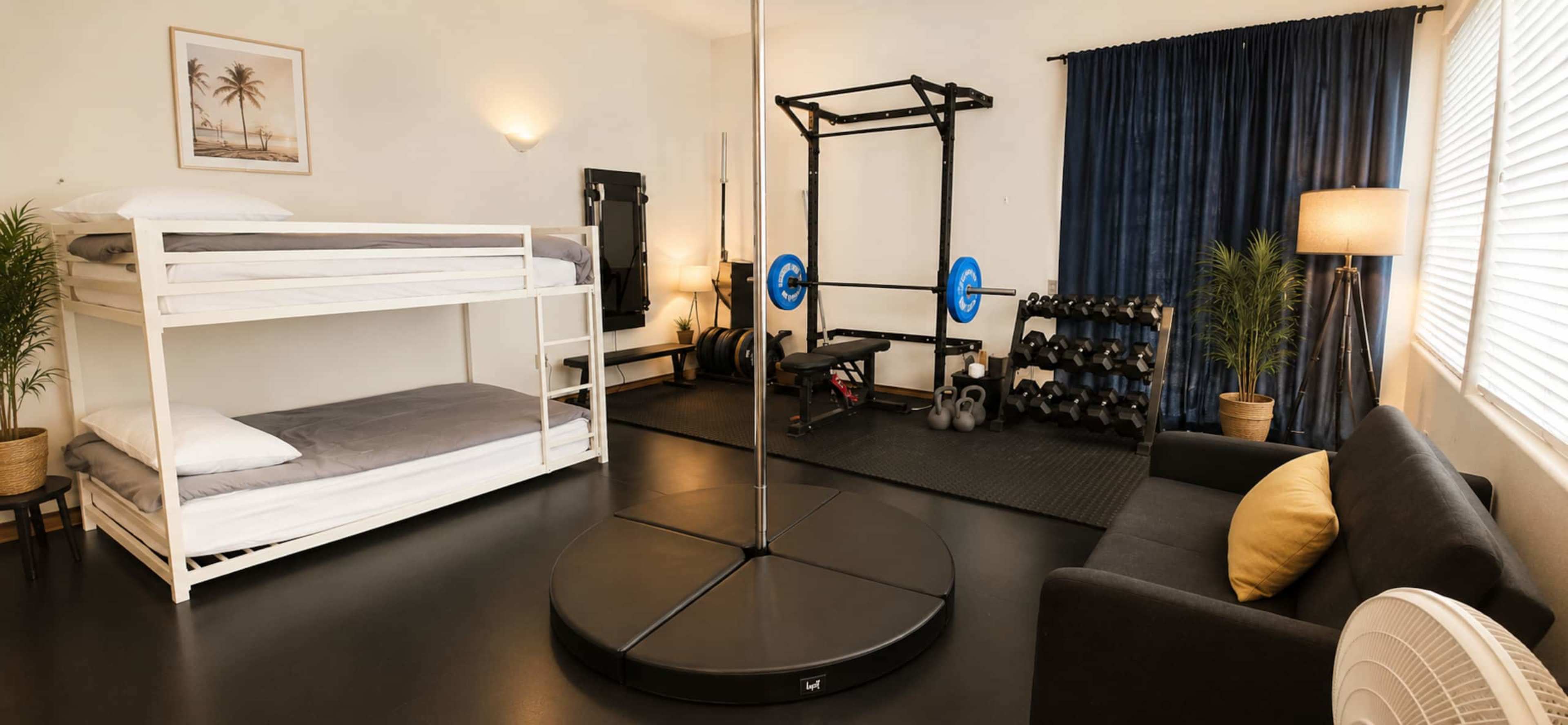 The image shows a studio room featuring a bunk bed, a weightlifting setup with a barbell, dumbbells, a pull-up bar, and a pole in the center, alongside a black couch and a lamp.