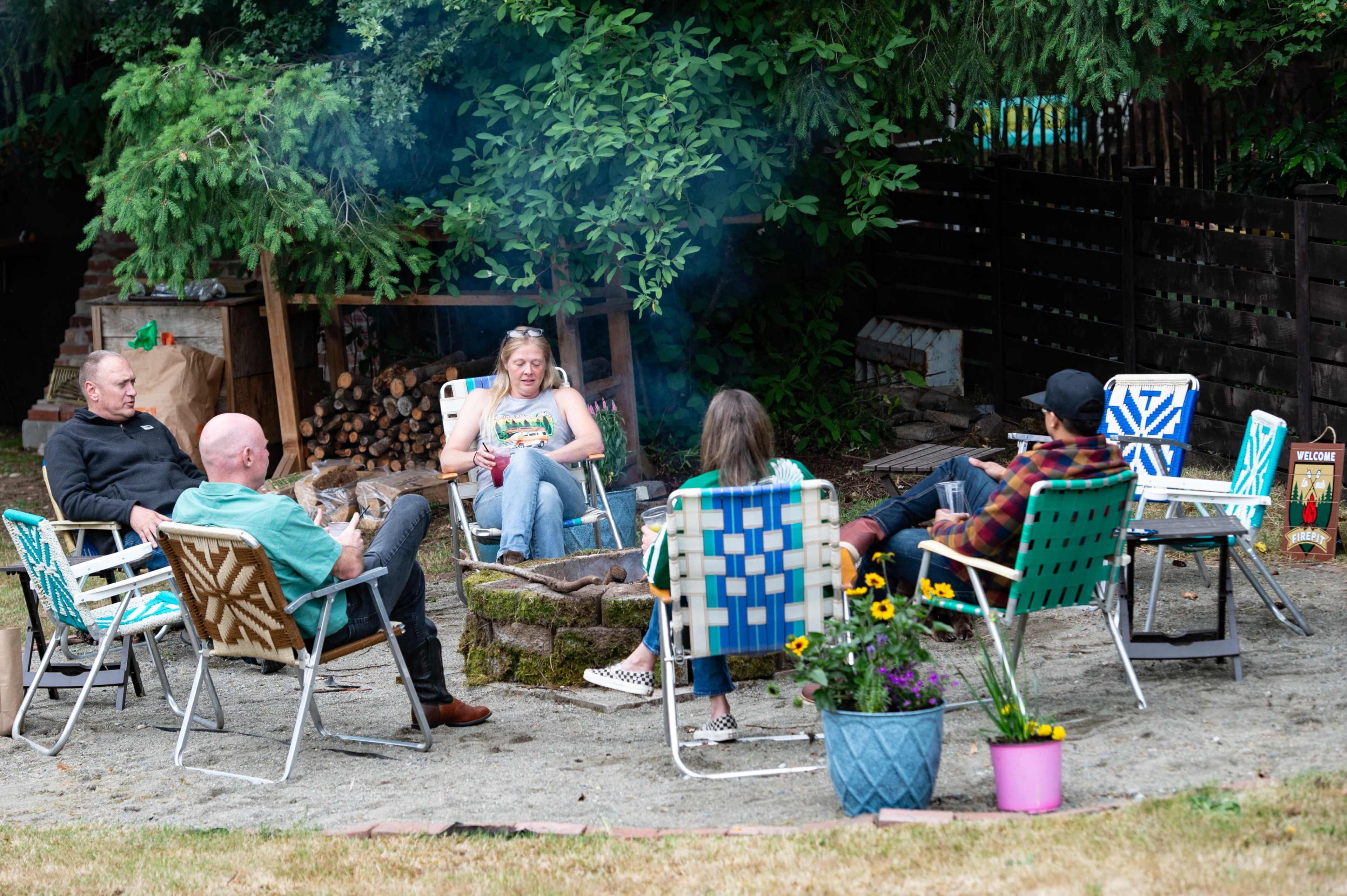 A group of six people sits around a fire pit in a backyard, surrounded by greenery and firewood stacked in the background.