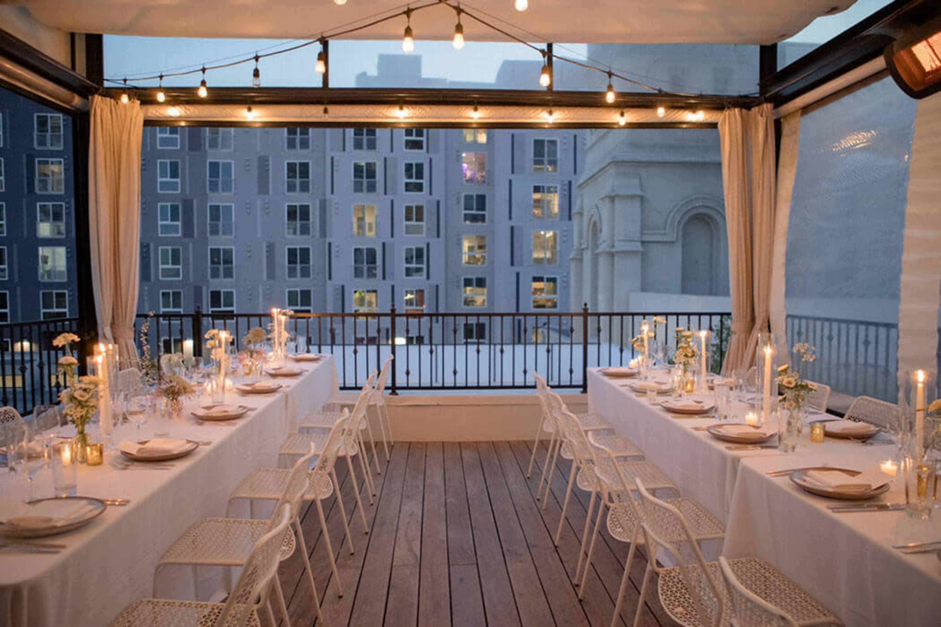 A decorated dining space with two long tables set for a meal is situated on a balcony overlooking a cityscape, illuminated by string lights and candlelight.