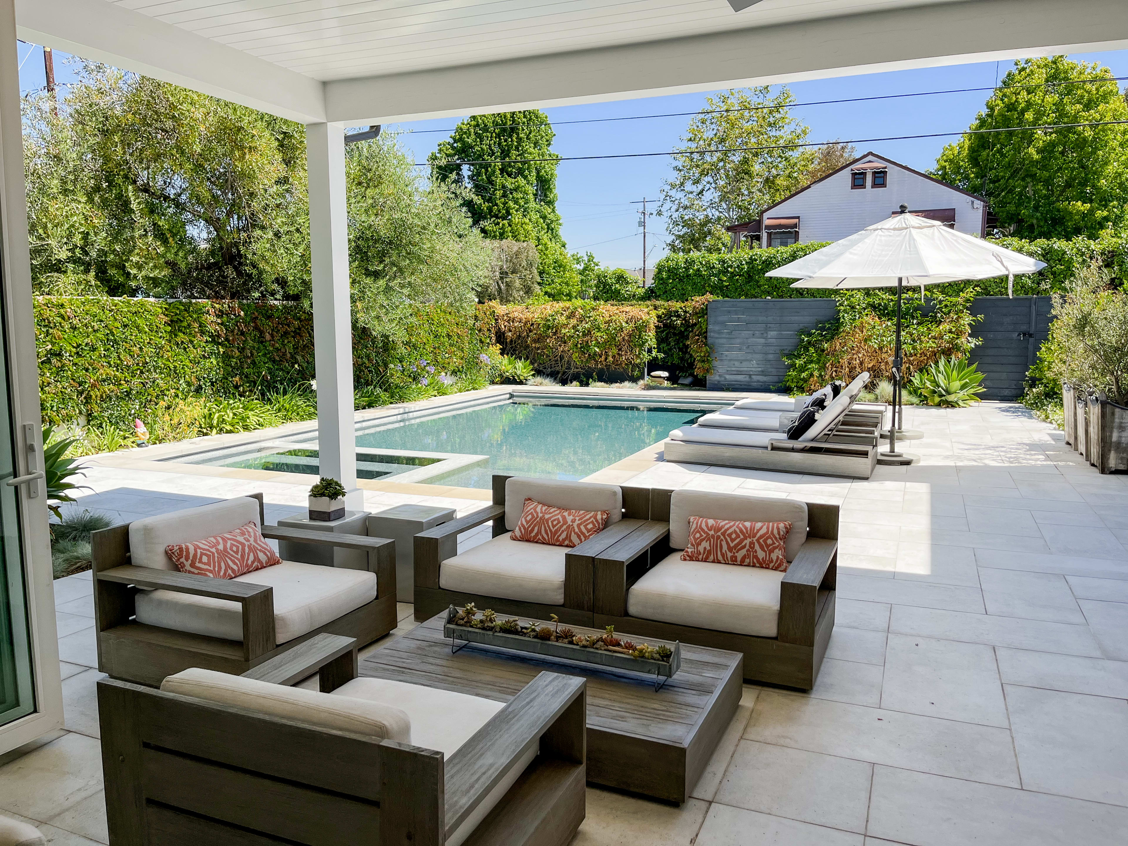 A modern outdoor patio features comfortable seating, a rectangular swimming pool, and a shaded umbrella area surrounded by lush greenery.