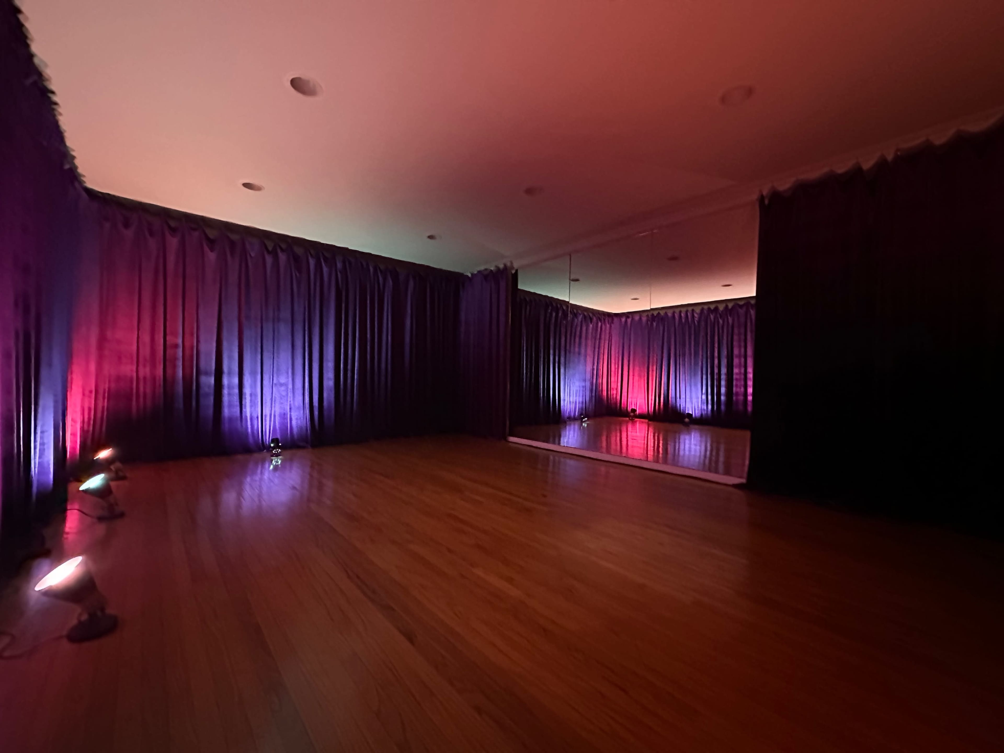 The image shows a dimly lit room with hardwood floors, featuring curtains and colored lights creating a moody atmosphere.